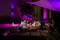 A couple sits relaxed in the evening in an illuminated outdoor lounge under a sunshade, surrounded by purple light.