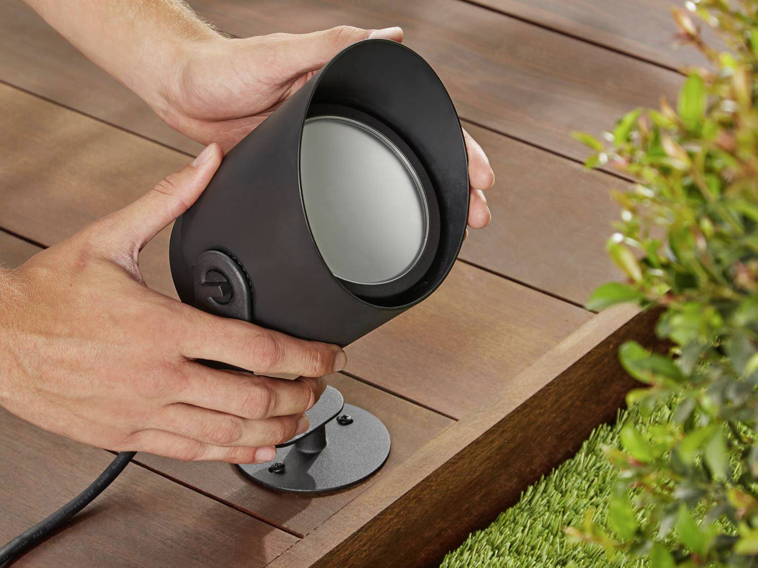 A person is installing a black outdoor light on a wooden decking, near an area with green plants.