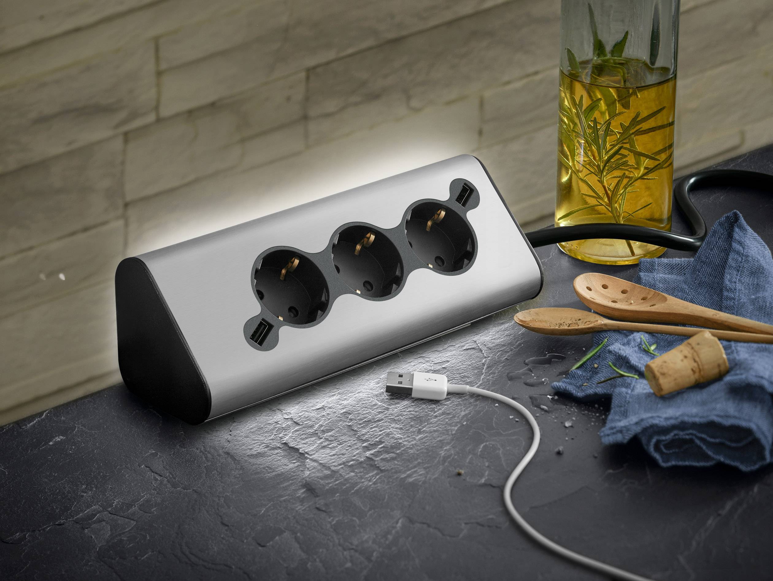 Multiple socket with three outlets on a kitchen worktop, surrounded by olive oil, wooden spoons and a USB cable.
