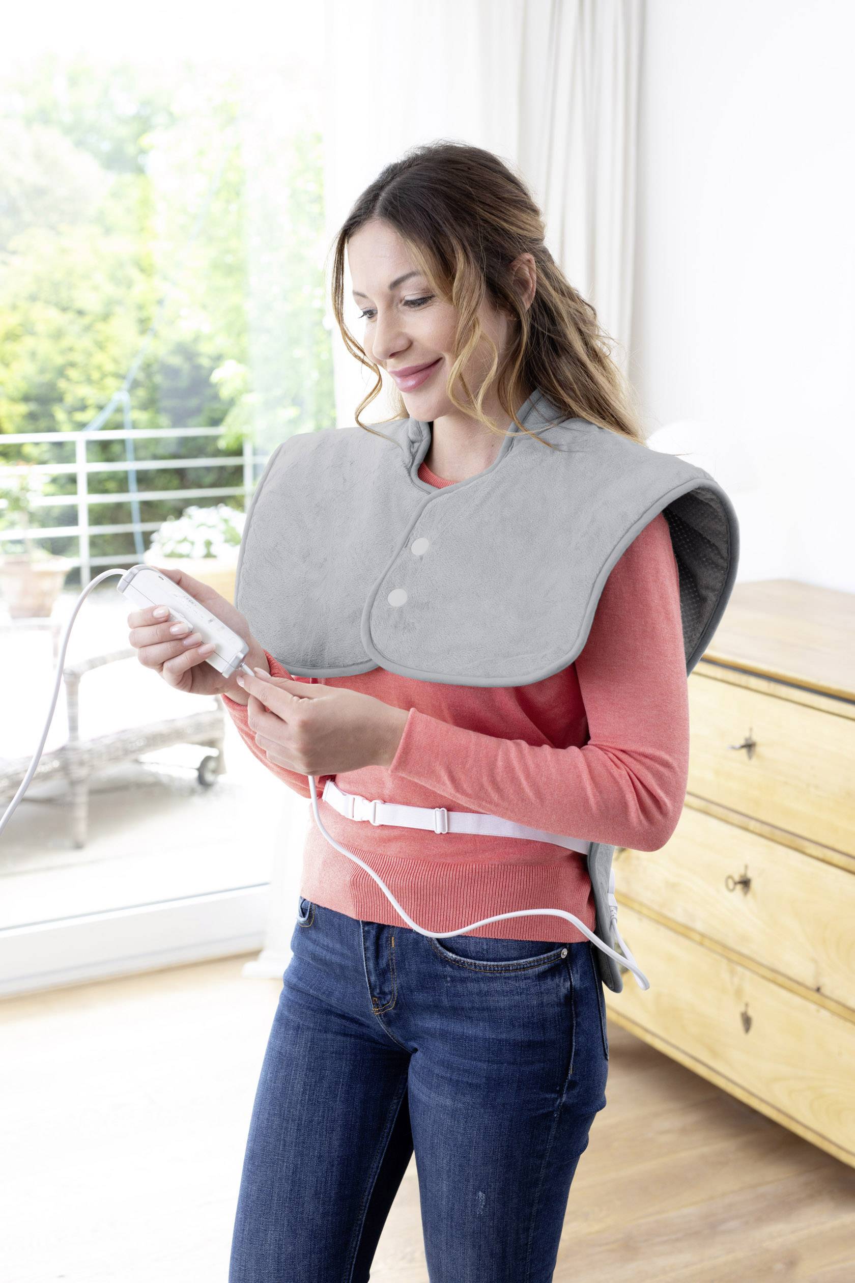Woman in living room wearing a grey shoulder heating blanket and holding a controller. Window with green view in the background.