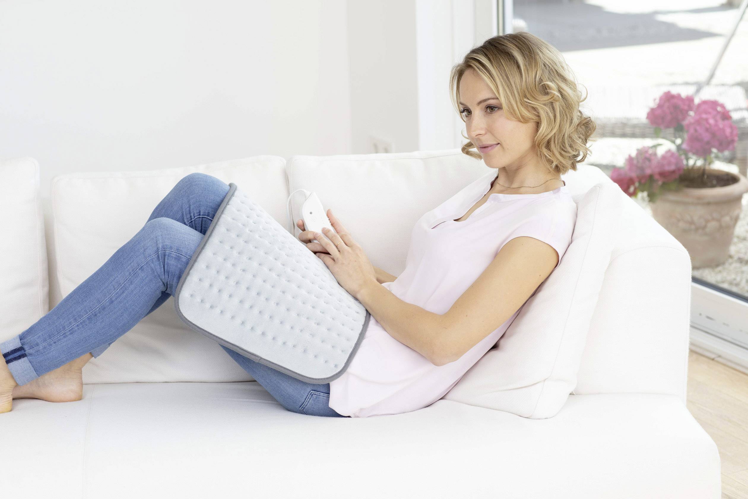 A woman sits relaxed on a sofa with a heated blanket on her legs and is holding a remote control. In the background, there are windows and flowers.