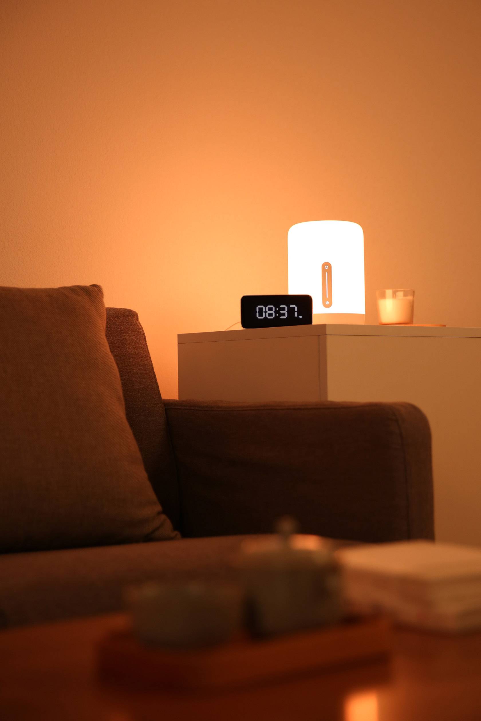 A clock on a bedside table shows 08:37 next to a glowing lamp in a warm, cosy living room.