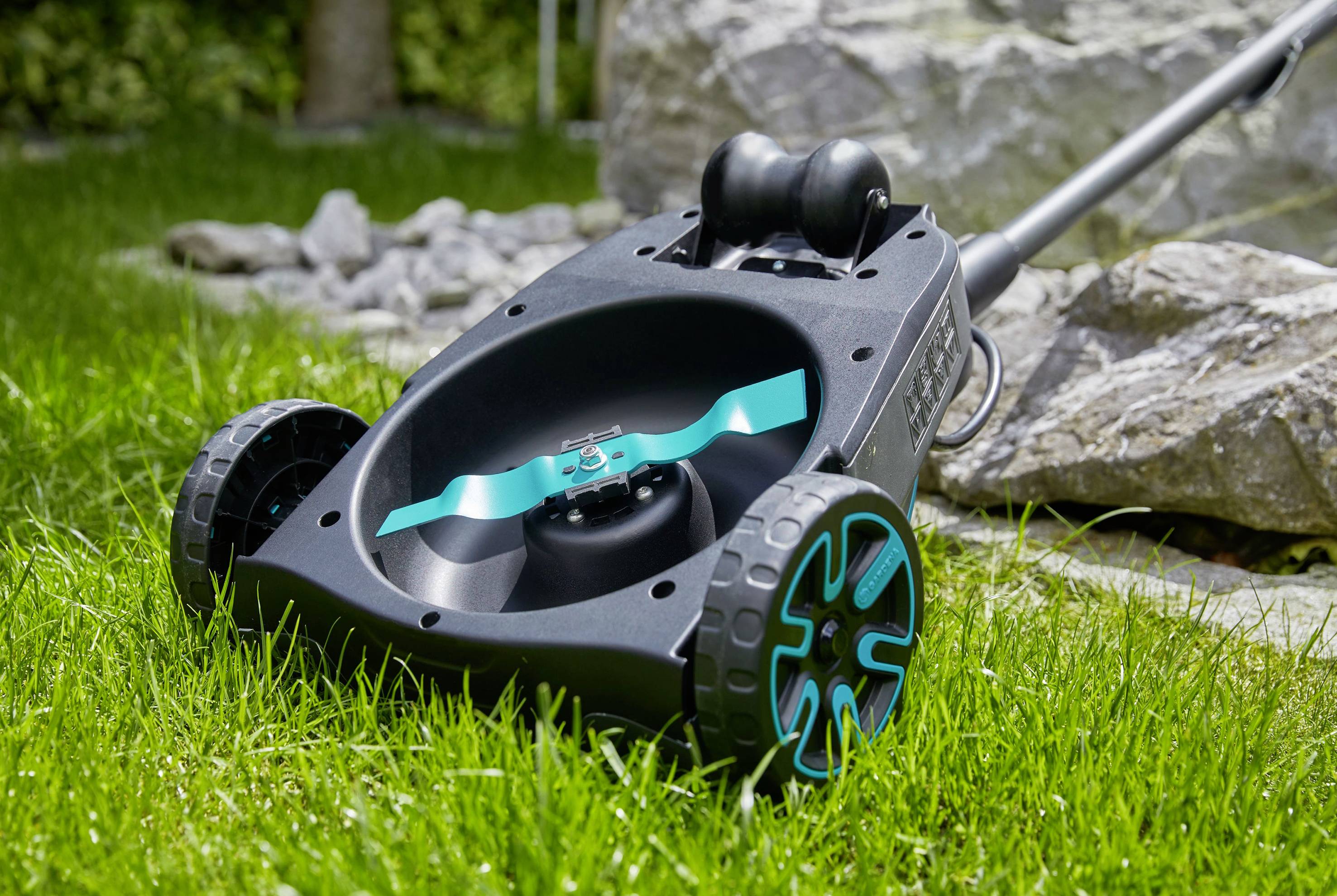 A lawnmower with a blue-black design sits on a green lawn. Stones can be seen in the background.
