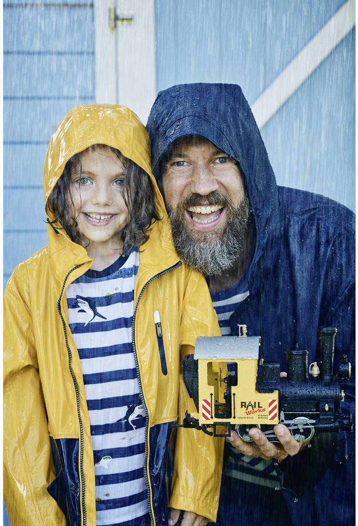 A cheerful adult and a child, both wearing raincoats, are standing in the rain. The adult is holding a small model train piece.