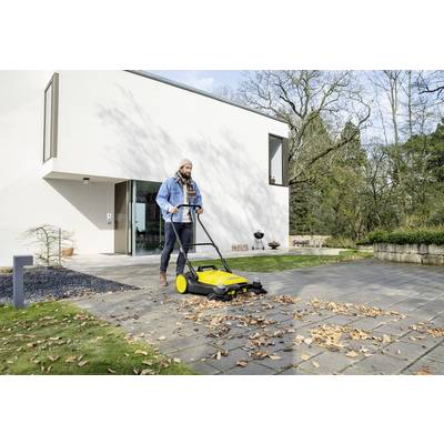 A person uses a yellow push sweeper to clear leaves from a modern home's driveway, surrounded by trees and greenery.