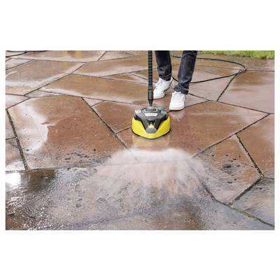 A person is using a yellow power washer to clean a red stone patio. Water sprays from the nozzle, removing dirt and brightening the stones.