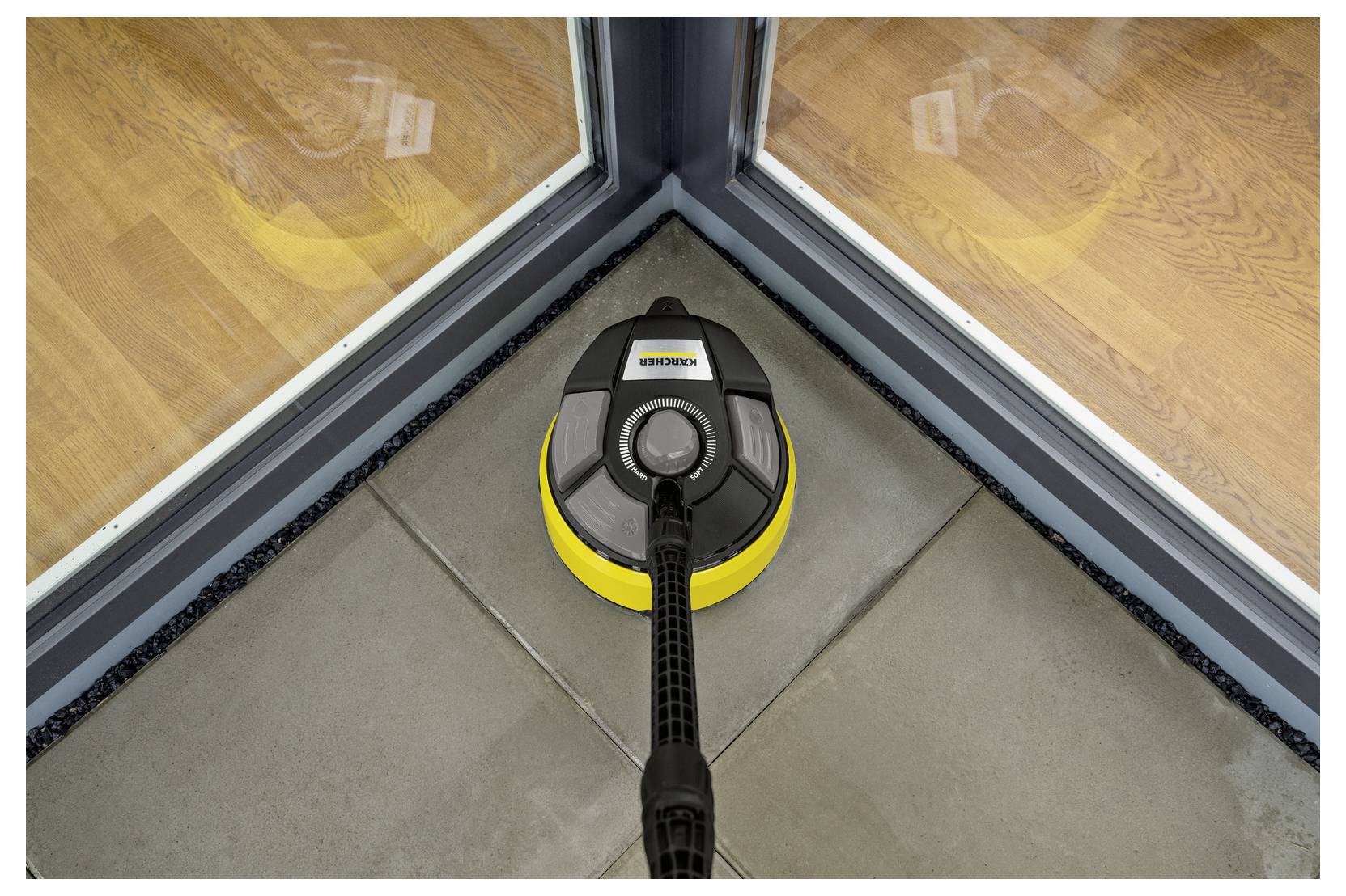 A floor cleaning device is shown cleaning a tile floor near the corner of a glass door, demonstrating its ability to clean edges effectively.