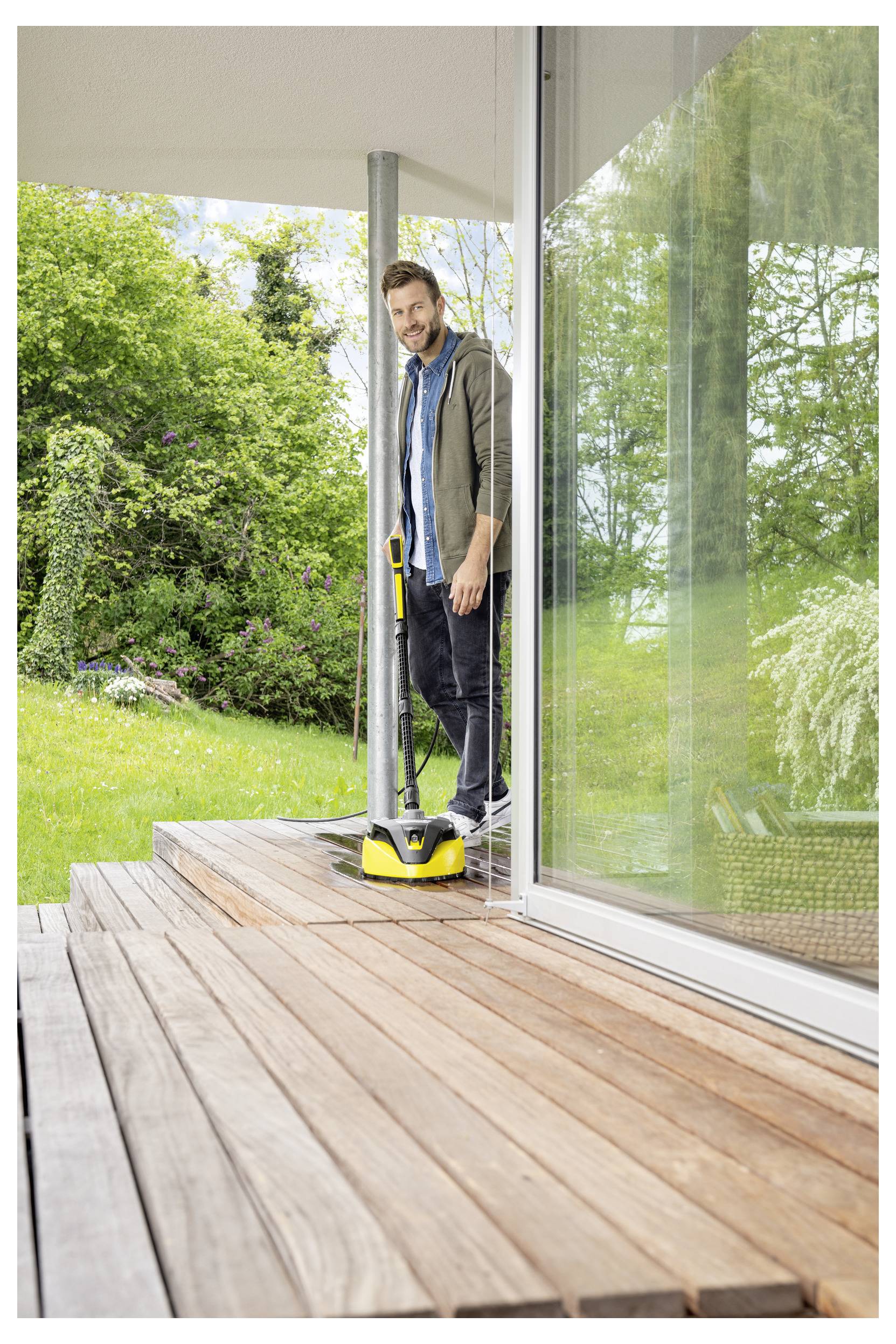 A person is using a yellow power washer to clean a wooden deck outside a modern house, surrounded by greenery and a glass door.