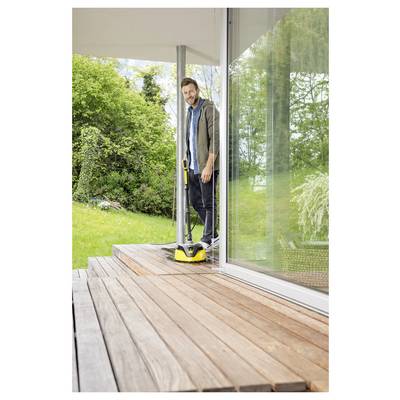 A person is using a yellow power washer to clean a wooden deck outside a modern house, surrounded by greenery and a glass door.