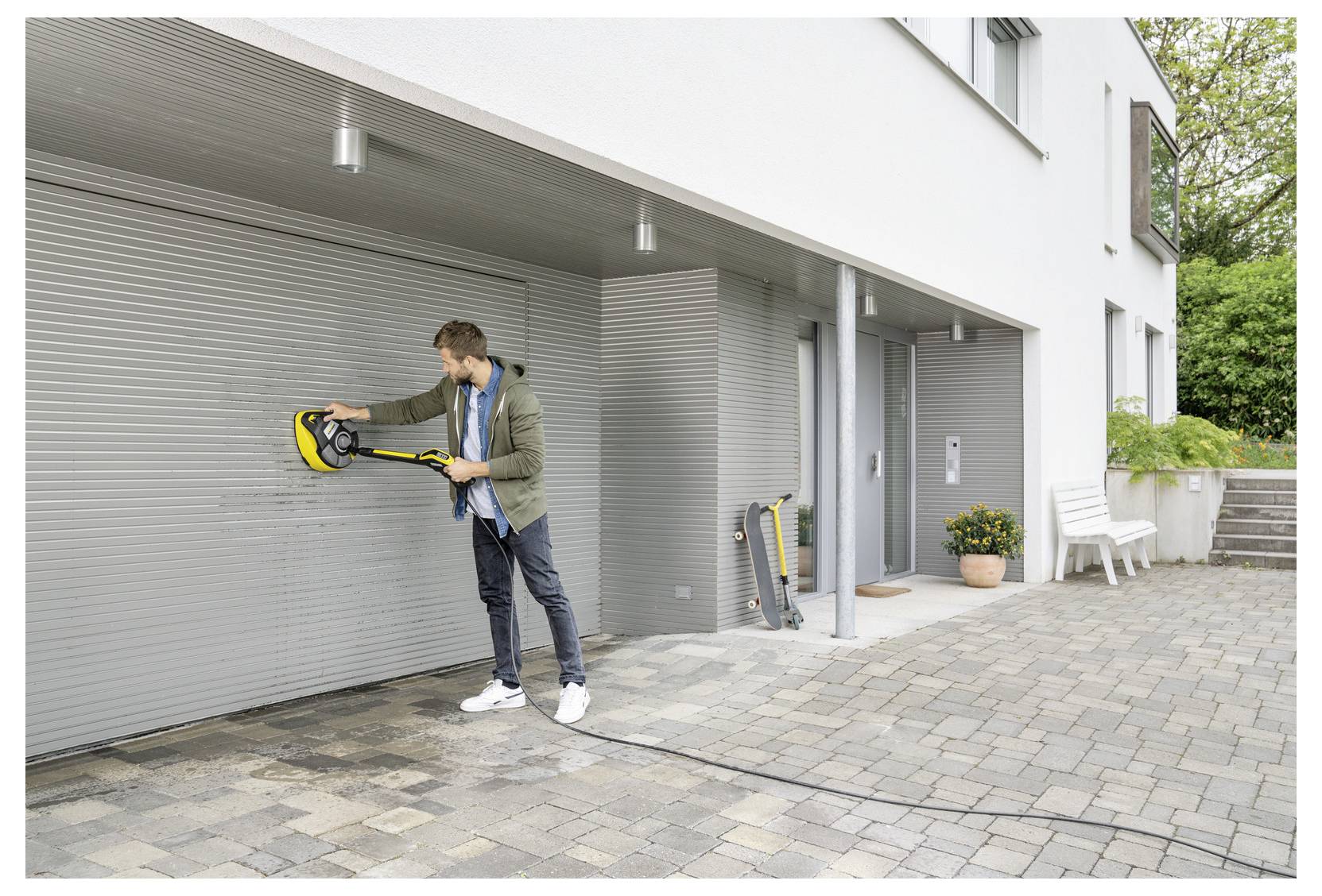 A person cleans a white garage door using a pressure washer on a paved driveway outside a modern house.