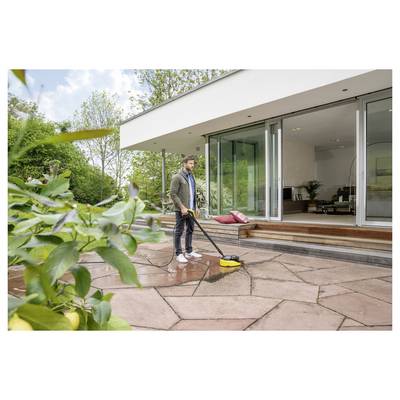 A person cleans a large stone patio with a pressure washer outside a modern house with large glass doors. Bushes and trees surround the area.