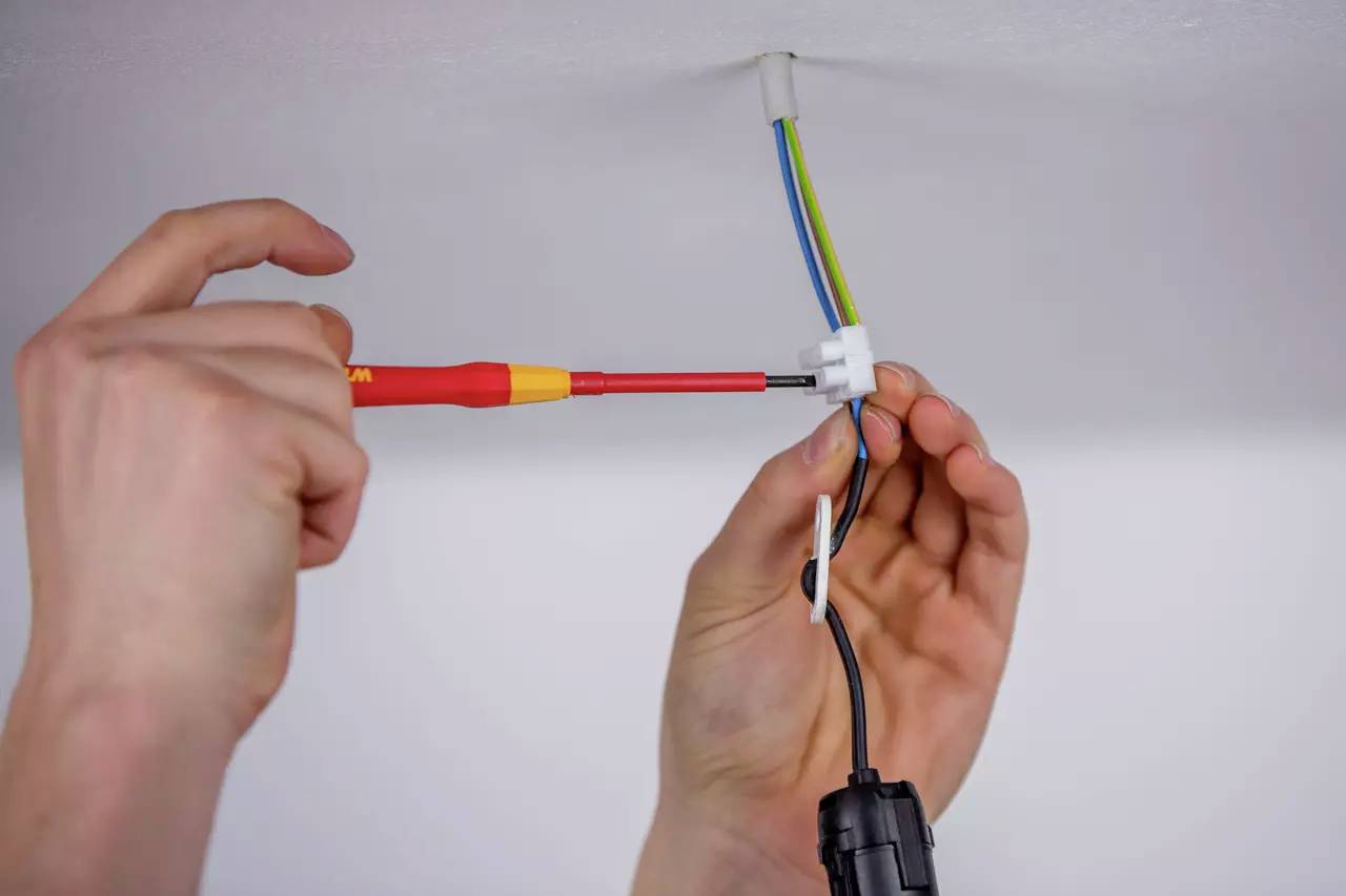A person is holding a screwdriver and connecting cables in an electrical socket on the ceiling.