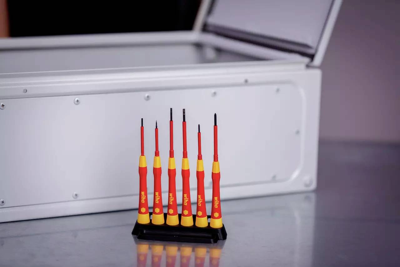 Six red screwdrivers in a stand in front of an open, grey metal toolbox on a table.