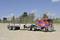 Lorry with a red cab and a flat trailer, unloaded, parked on sandy ground with trees in the background. Clear sky.