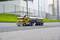 A yellow model lorry with a tanker trailer is parked on a road in front of a green lawn and an industrial backdrop.