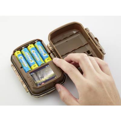 Hand opens brown camera housing with inserted AA batteries. Inside, the LCD screen displays a photo of a deer in the forest.