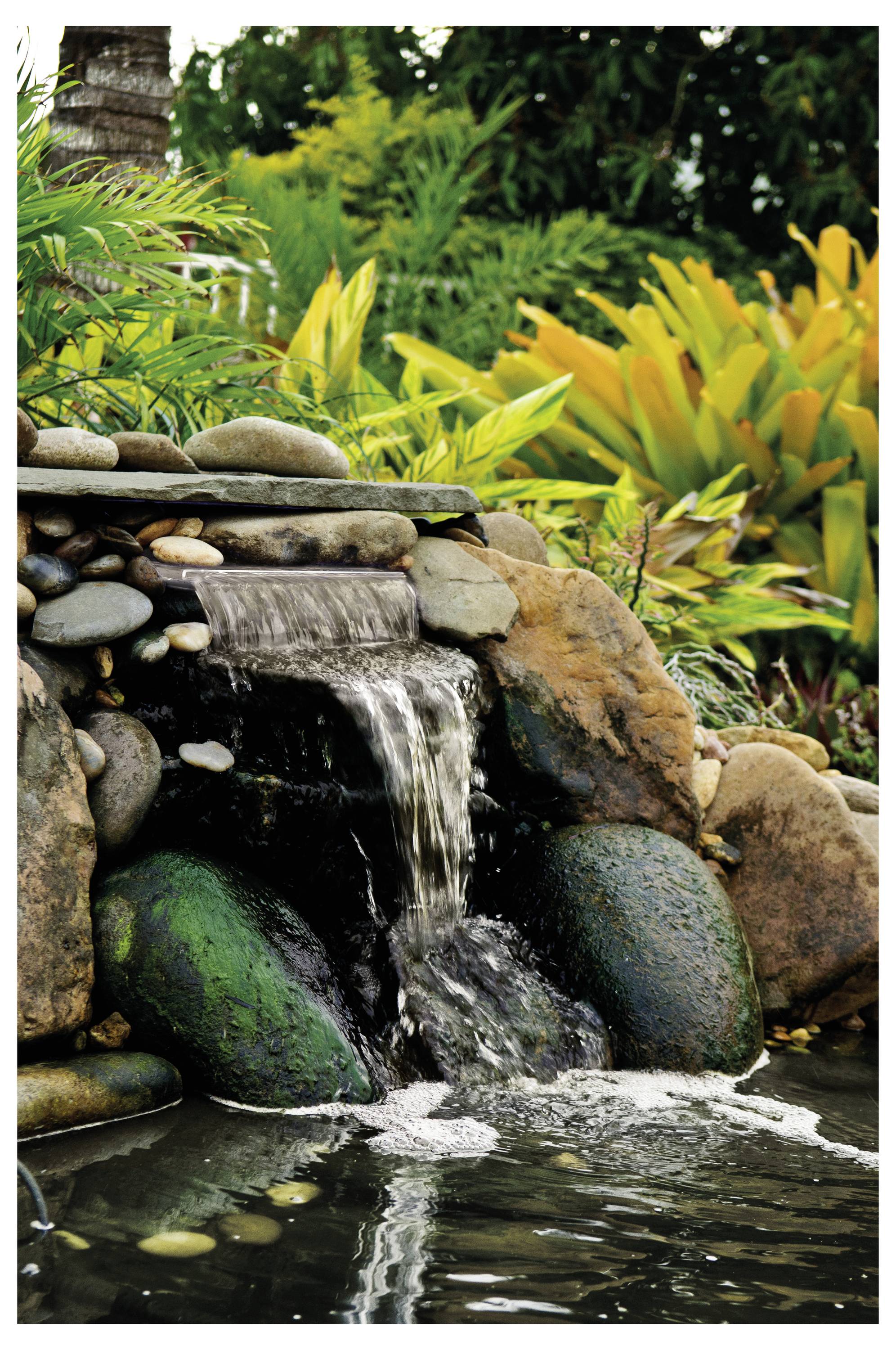 A small waterfall flows over rocks into a pond, surrounded by lush green and yellow foliage, creating a tranquil garden scene.