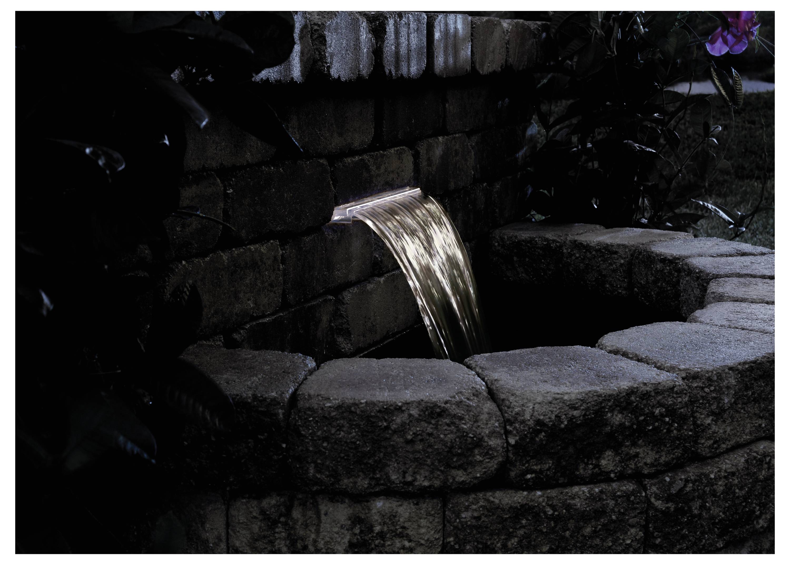 'A small waterfall flows from a stone wall into a circular stone basin, surrounded by leaves, in a dark, serene setting.'