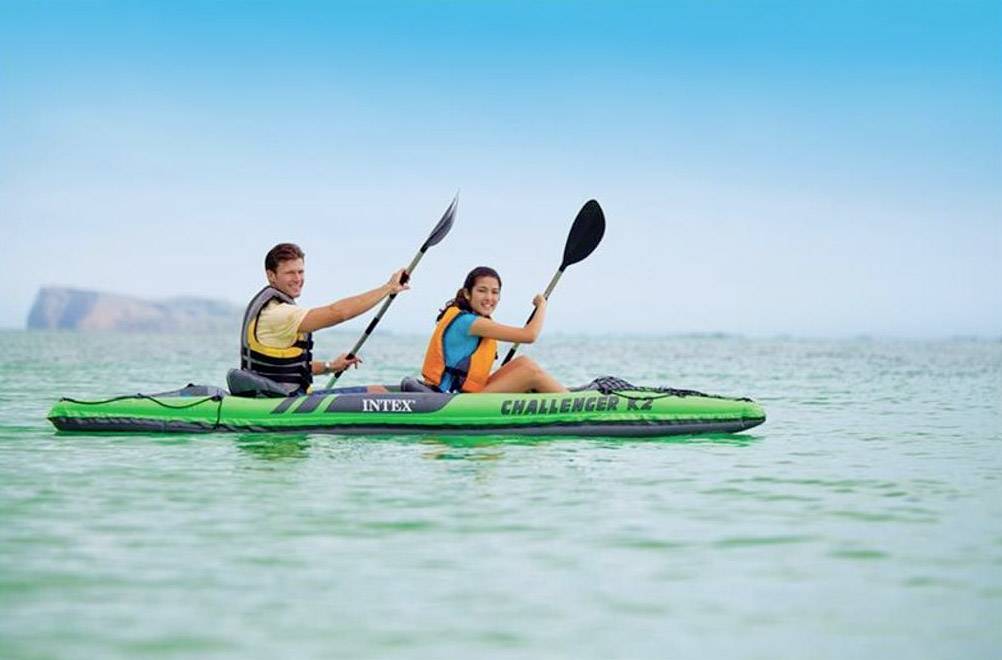 Two people are paddling in a green kayak on a calm, sunny lake. Both are wearing life jackets and appear cheerful.