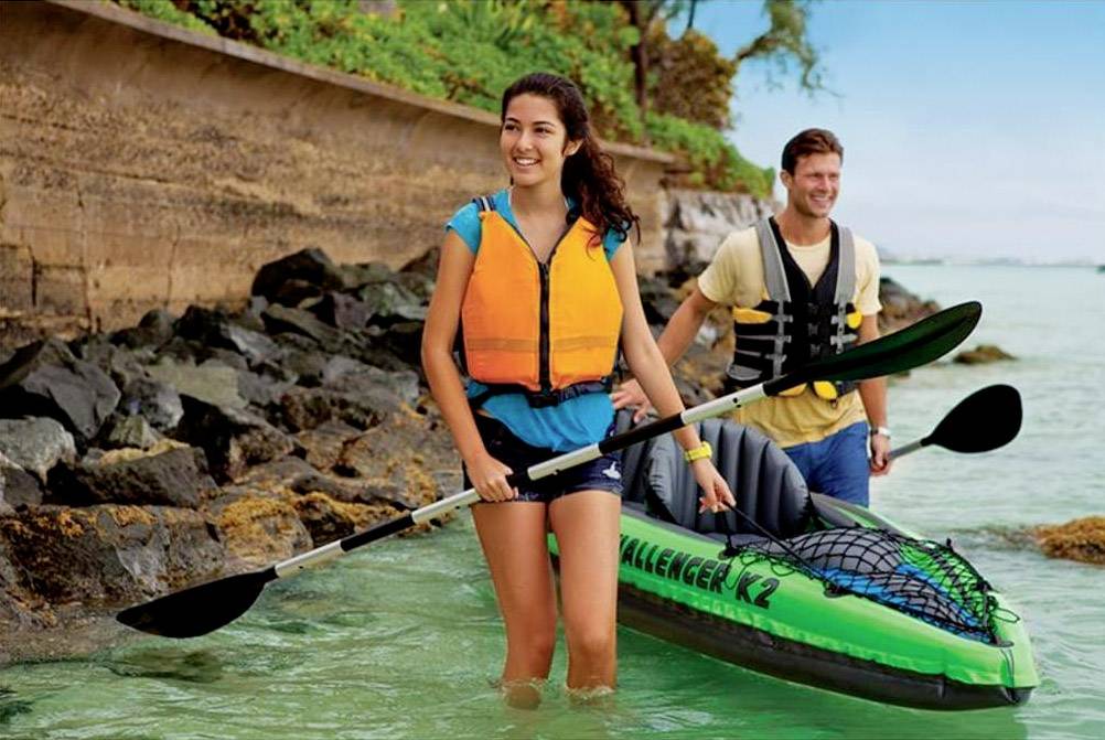 Two people in life jackets are standing smiling in shallow water next to a green kayak. A stone wall is visible in the background.