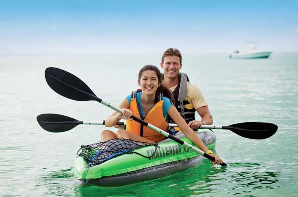 Two people are smiling and paddling in a kayak on calm blue water, both wearing life jackets.