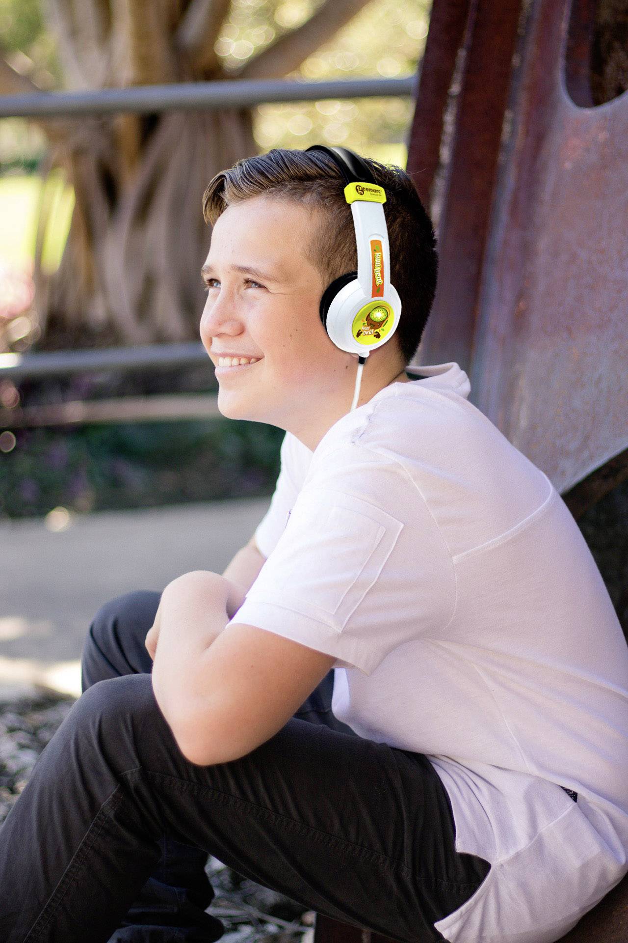 A boy is sitting outside wearing yellow headphones. He is looking up with a smile. Trees and a metal structure are visible in the background.