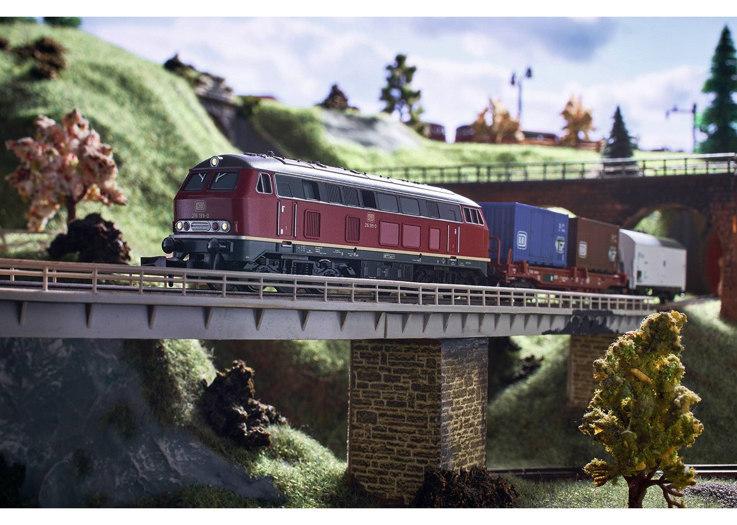 A red model locomotive pulls freight wagons across a bridge in a landscape with trees and hills.