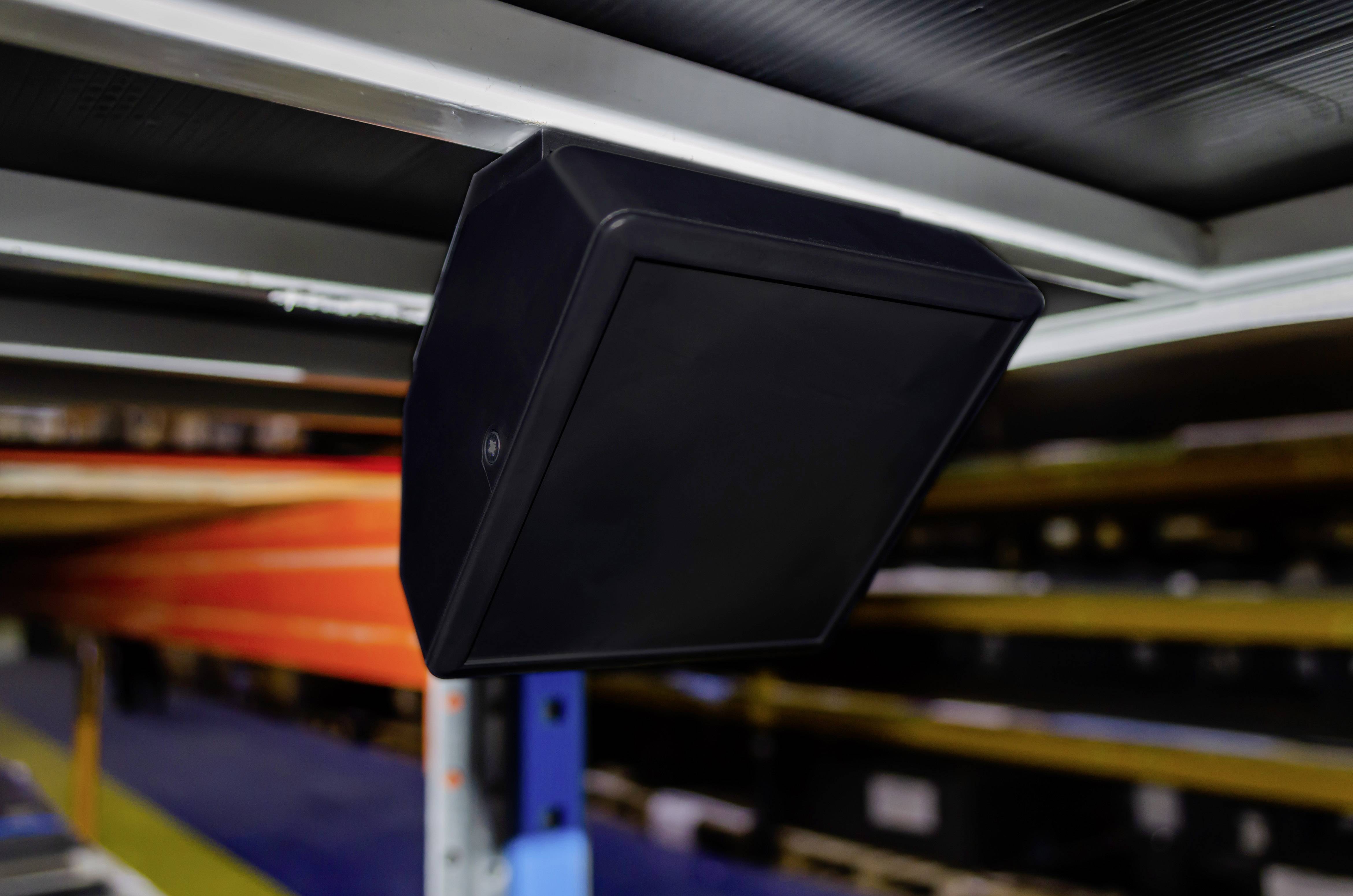 Black security camera mounted on the ceiling of a warehouse, aimed at shelves to monitor the area.