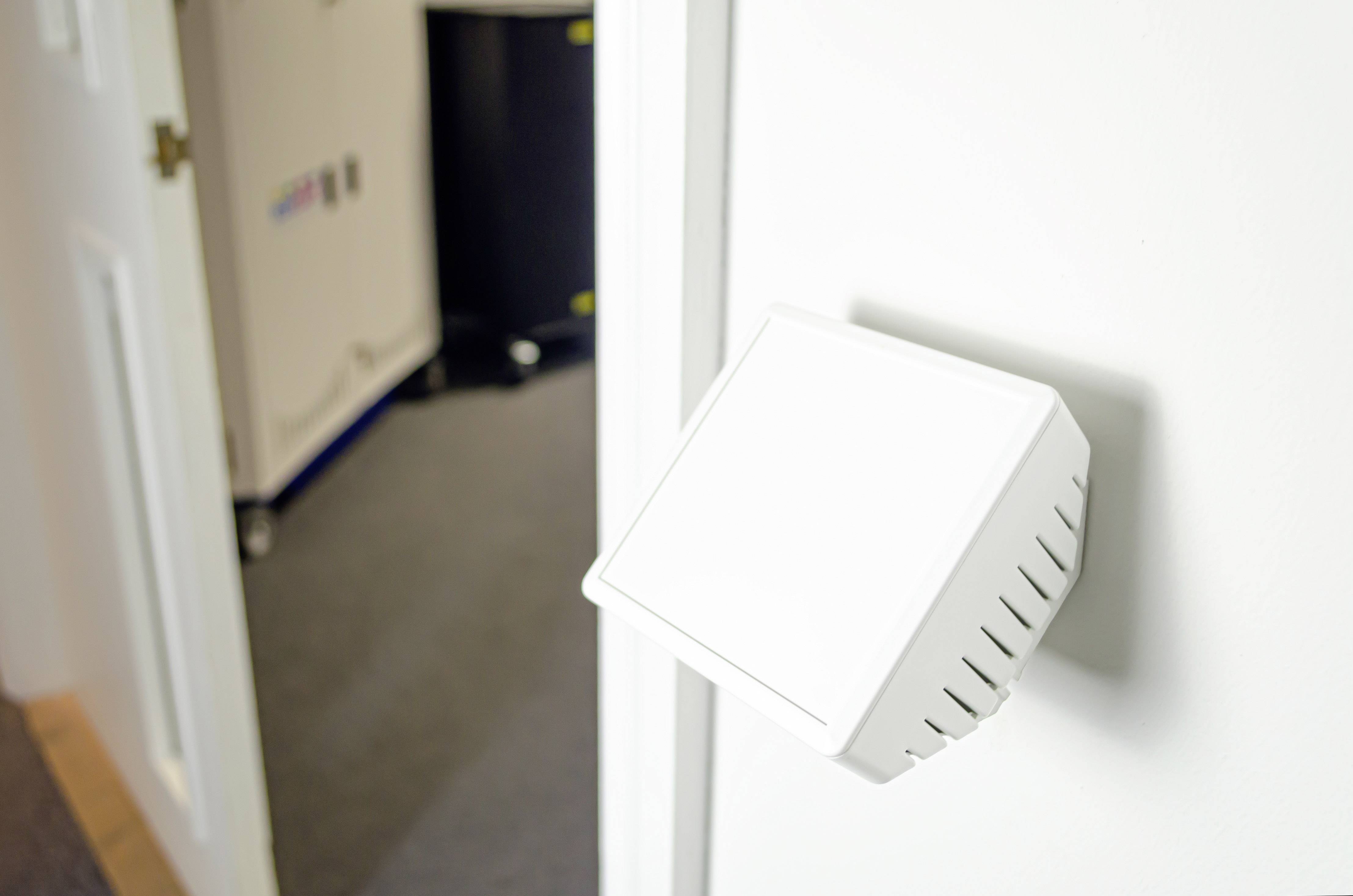 White, square device on a wall, next to a door, which is possibly a sensor or switch. A blurry room is in the background.