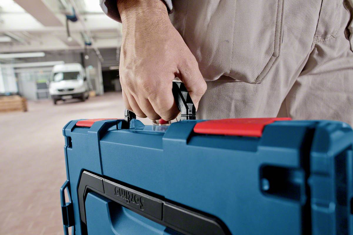 A person is carrying a blue toolbox in a workshop or garage, with a delivery van in the background.