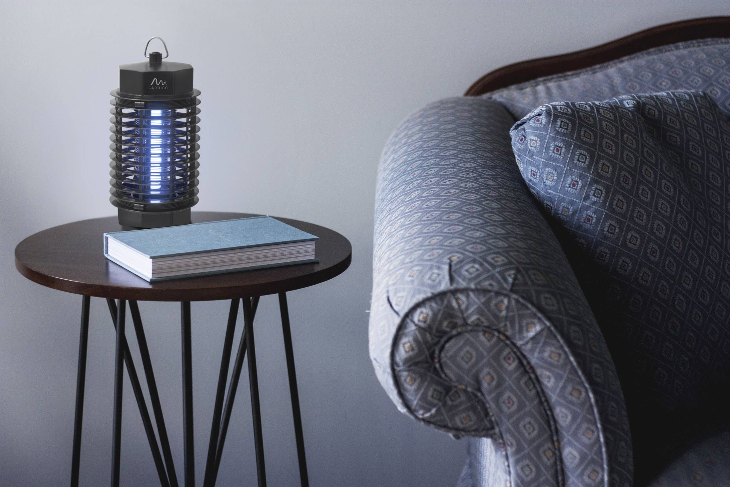 A sofa corner with a blue upholstered pattern. Next to it stands a table with a book and a switched-on insect lamp.
