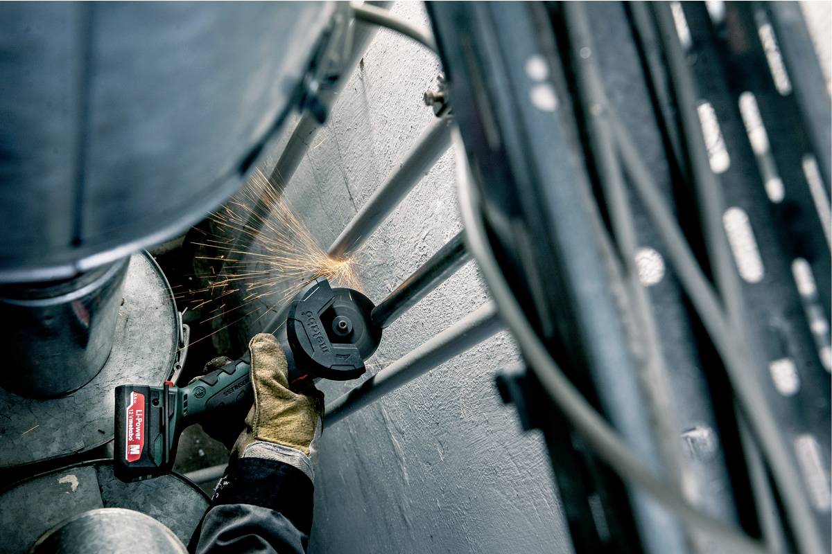 A person is grinding metal bars with an angle grinder, sparks flying. The scene depicts industrial work in a confined space.