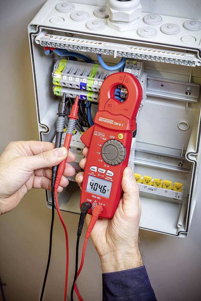 A person is holding a digital multimeter in front of an open fuse box and measuring a voltage of 404.6 volts.