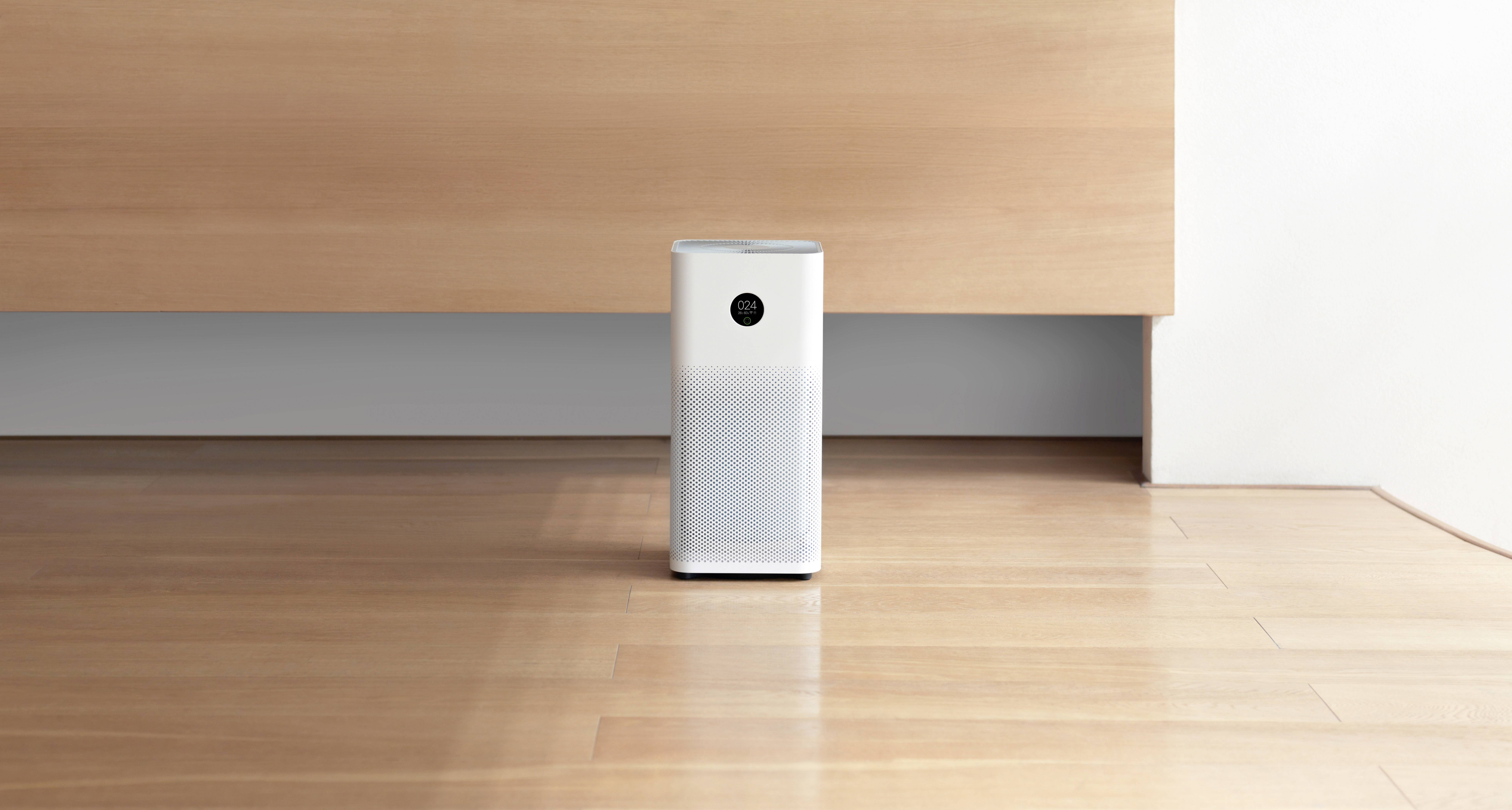 A white air purifier stands on a wooden floor in front of a light brown wall.