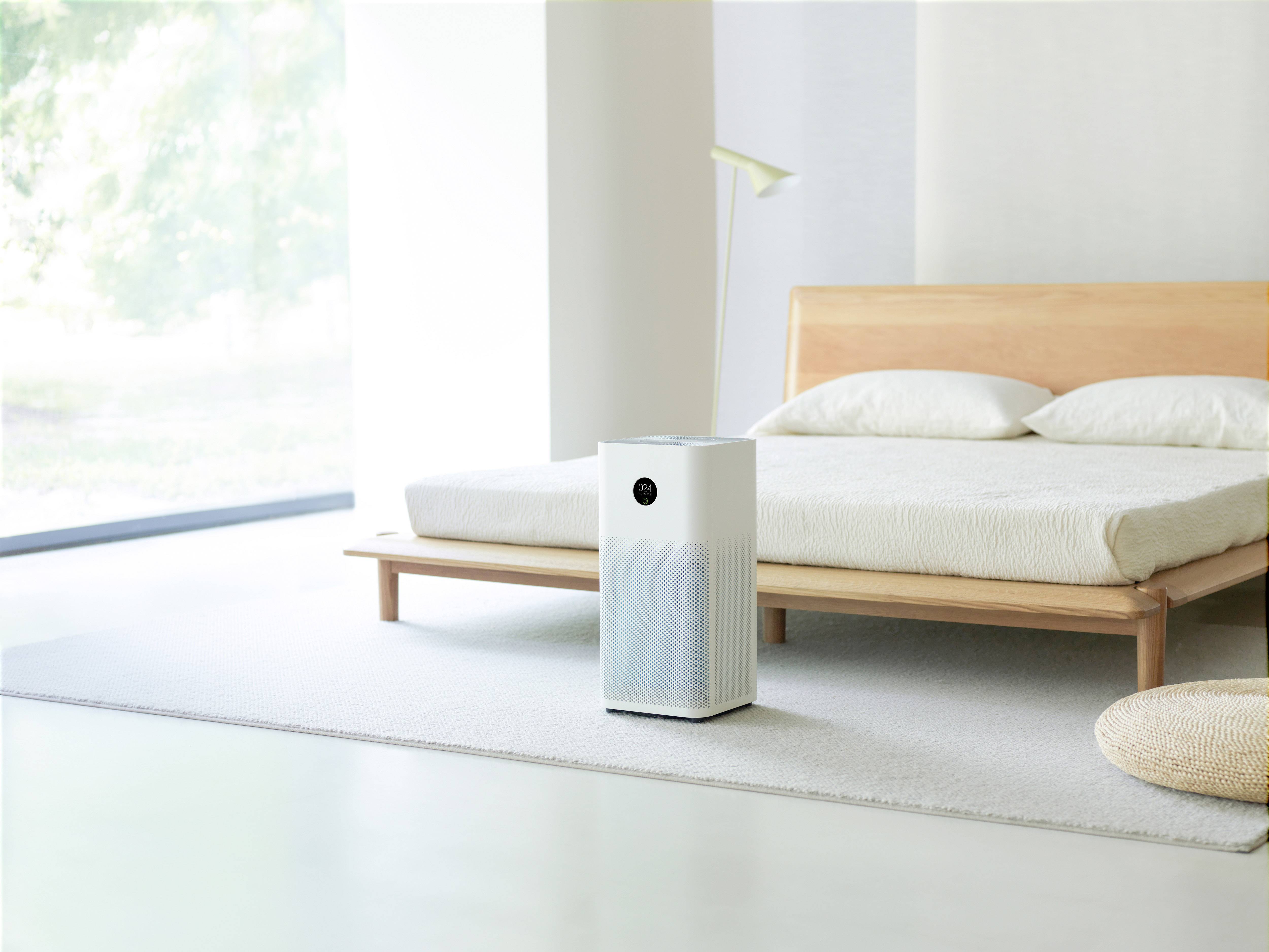 An air purifier stands in a minimally furnished bedroom next to a bed with a wooden frame and a white lamp.