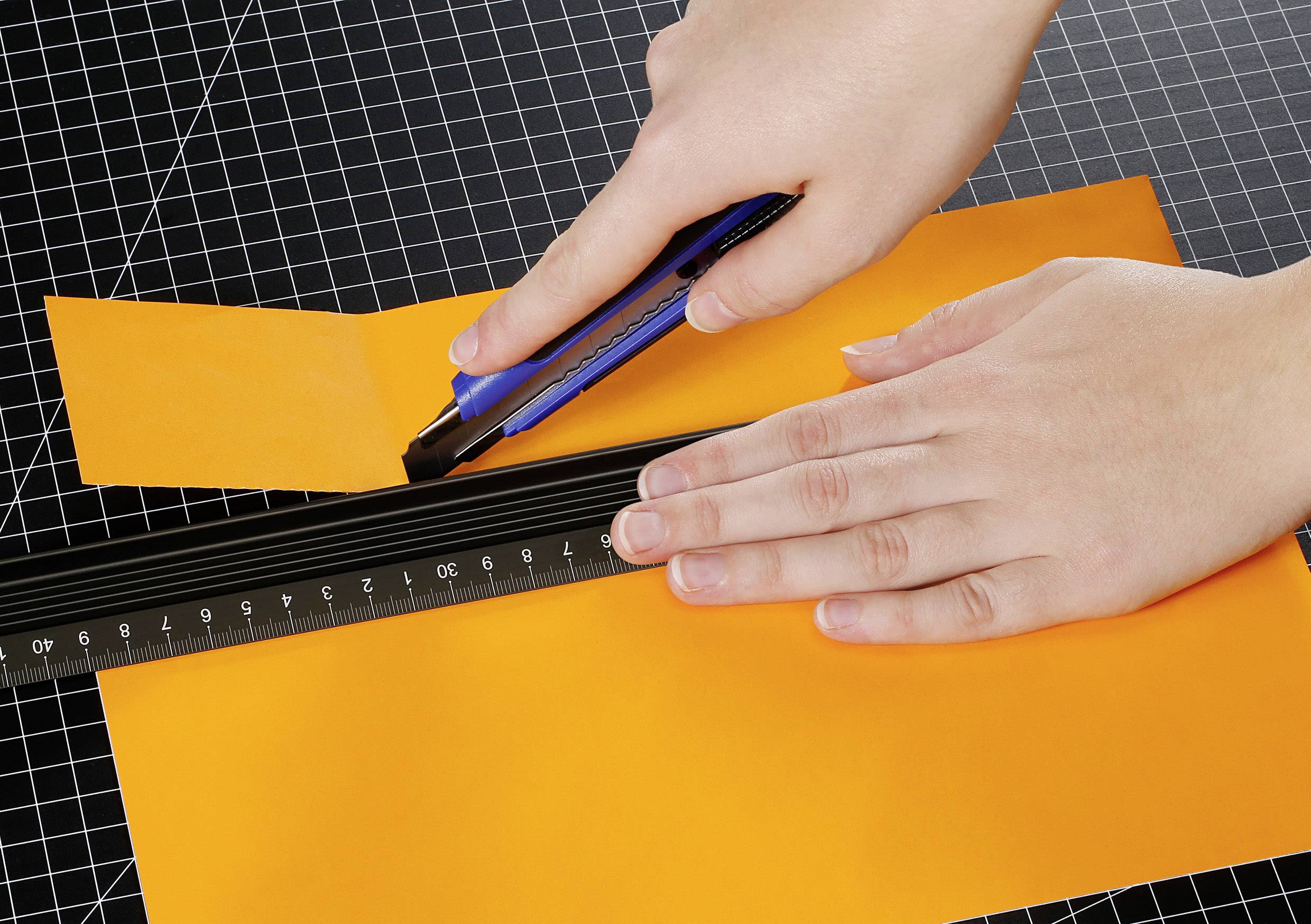 A person is cutting an orange piece of paper with a blue craft knife and a ruler on a chequered background.