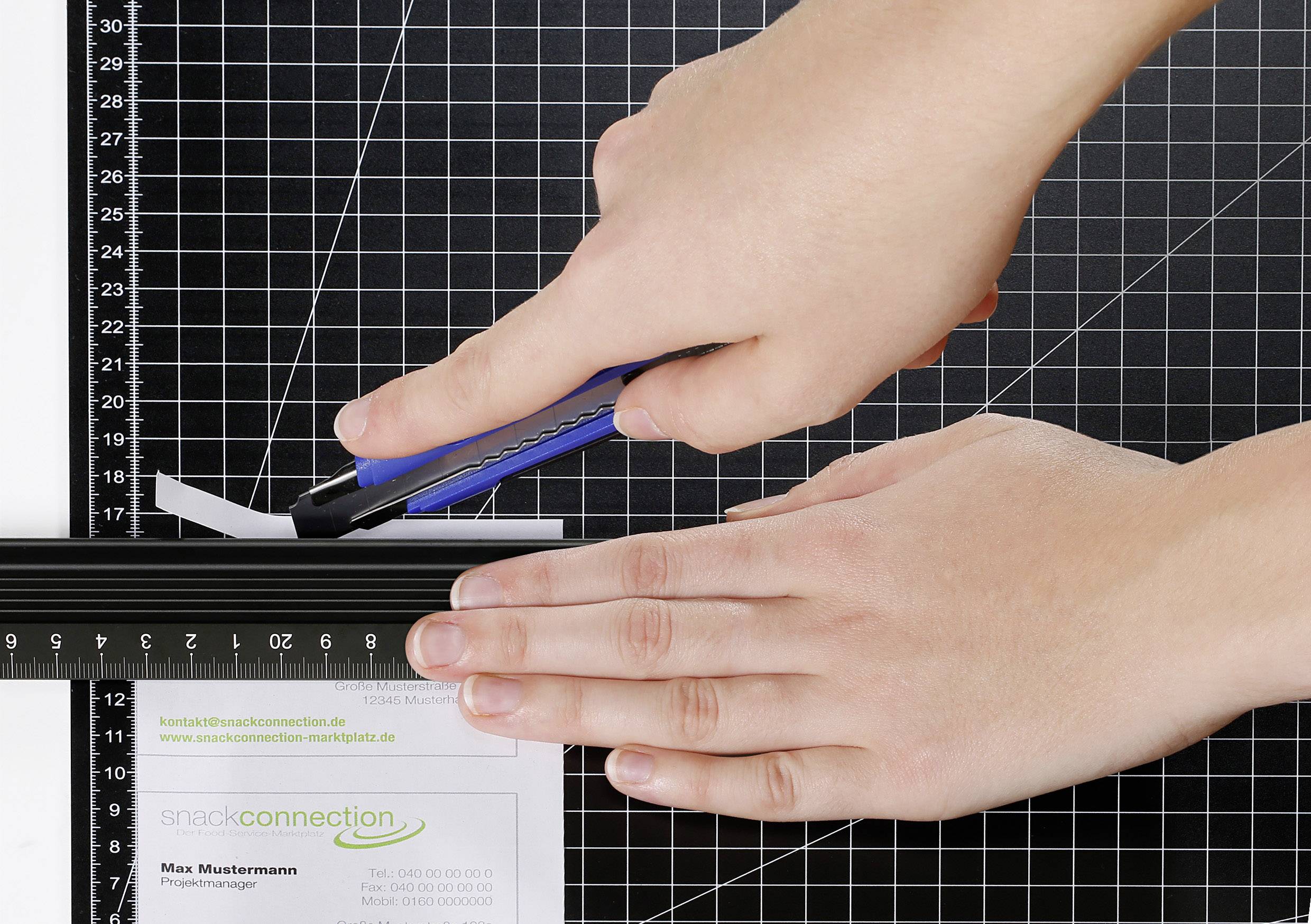 A hand is holding a blue craft knife and cutting a piece of paper on a chequered cutting board along a ruler.