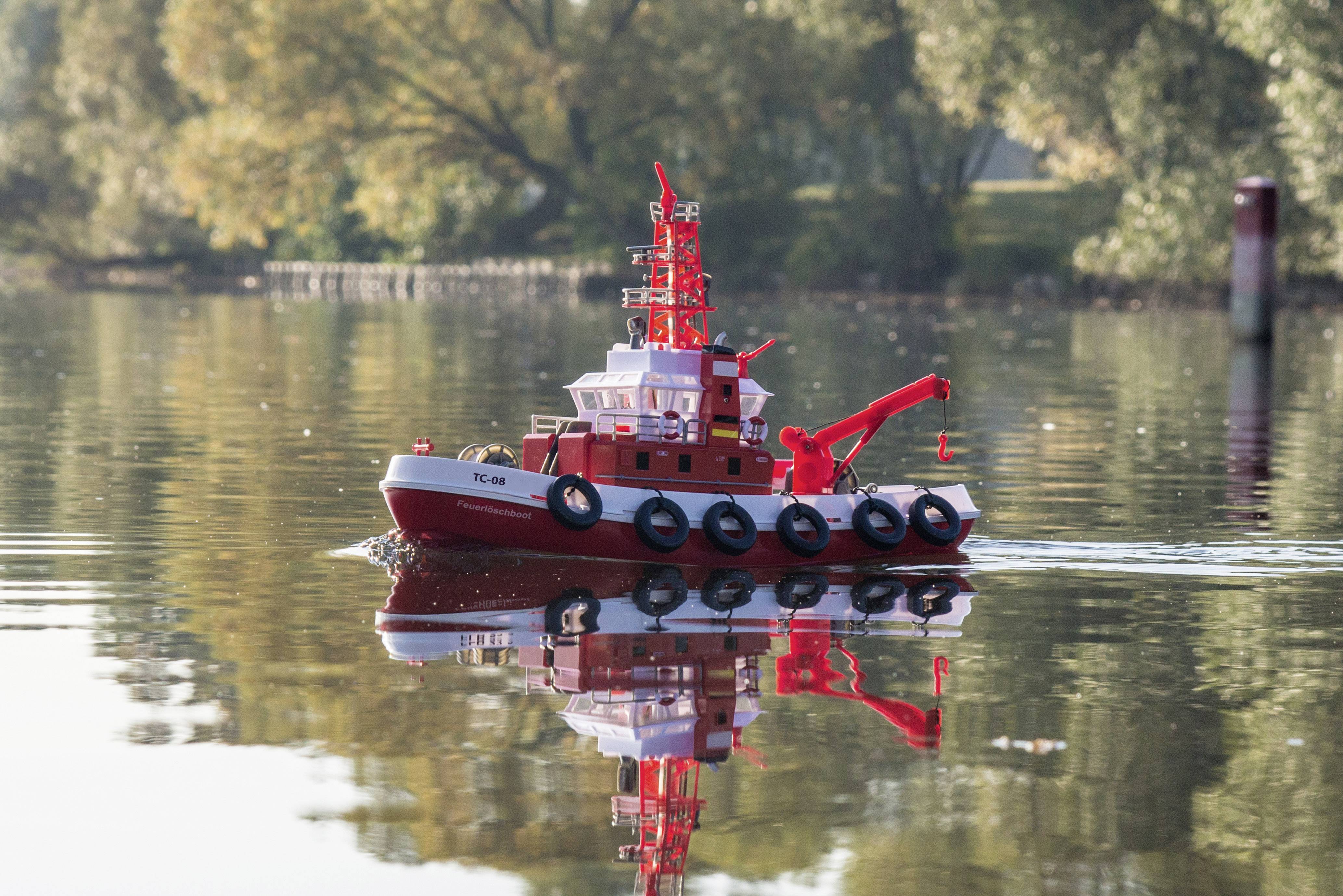 A small model of a red tug boat is floating on a calm lake. Trees and a wooded shoreline can be seen in the background.