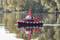 A small model of a red tug boat is floating on a calm lake. Trees and a wooded shoreline can be seen in the background.