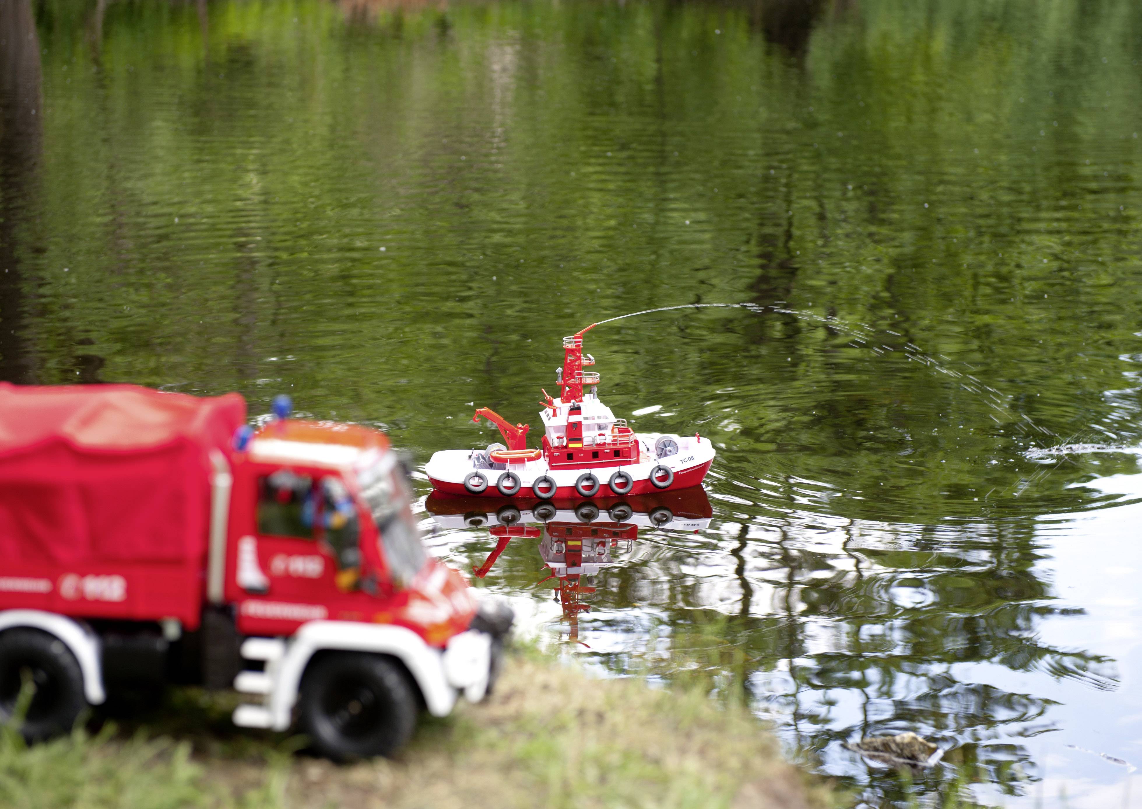 A small red fire engine and boat toy are in action in a pond. The boat is spraying water while the vehicle stands on the bank.