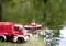 A small red fire engine and boat toy are in action in a pond. The boat is spraying water while the vehicle stands on the bank.