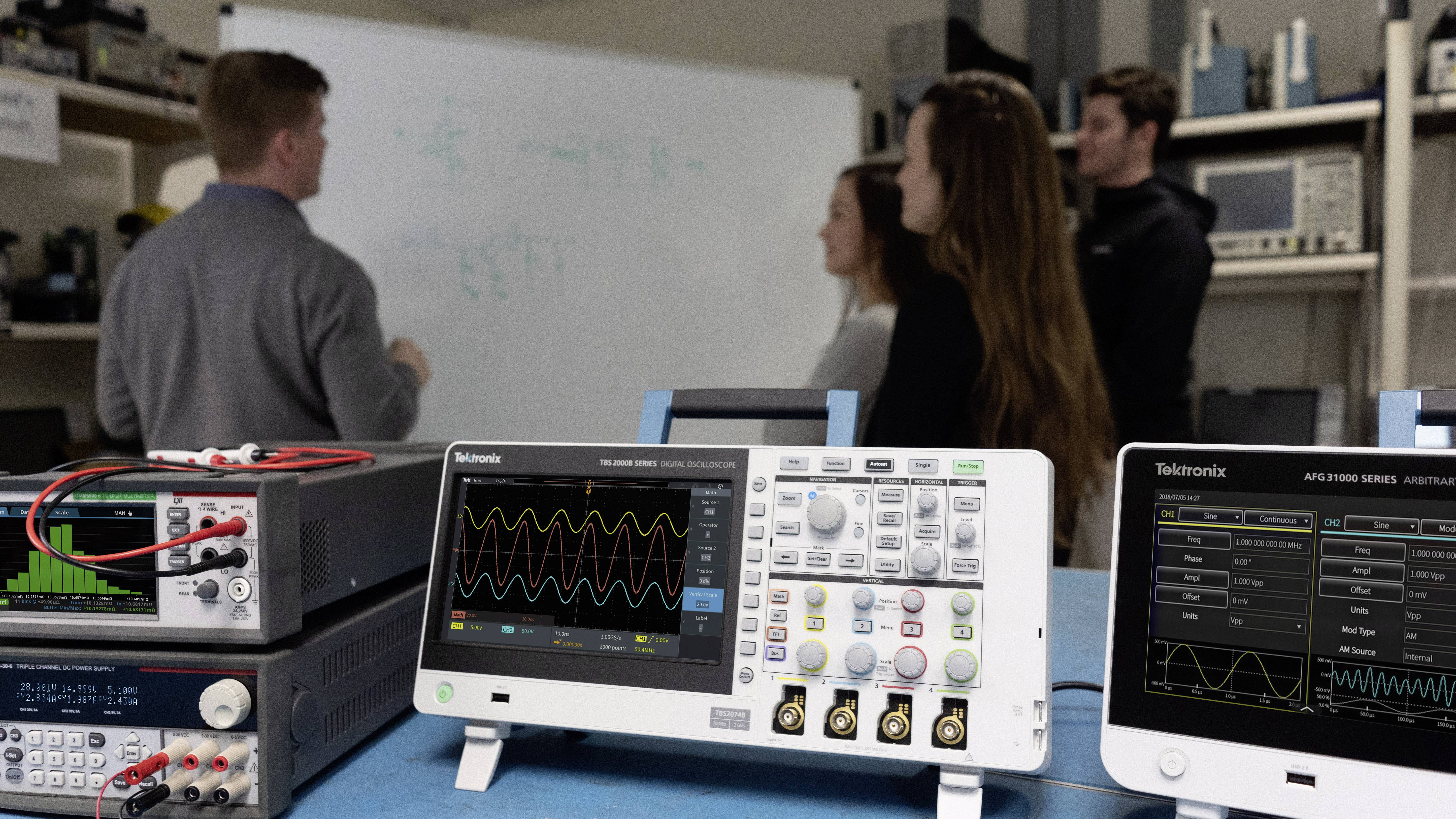 Four people are discussing an electronic circuit diagram on a whiteboard. Oscilloscopes are prominent in the foreground.