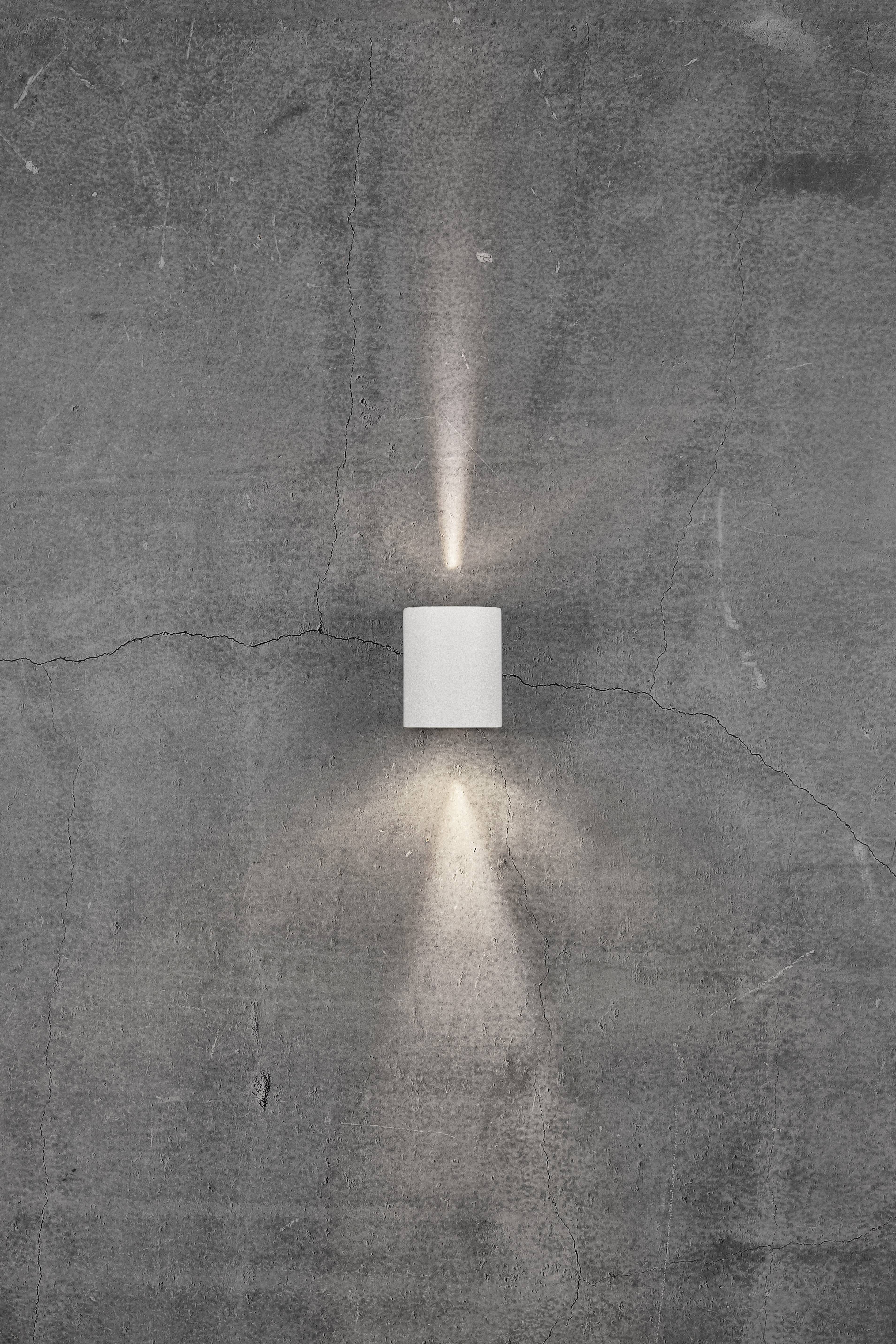 A white wall light on a grey, textured concrete wall emits light rays upwards and downwards.