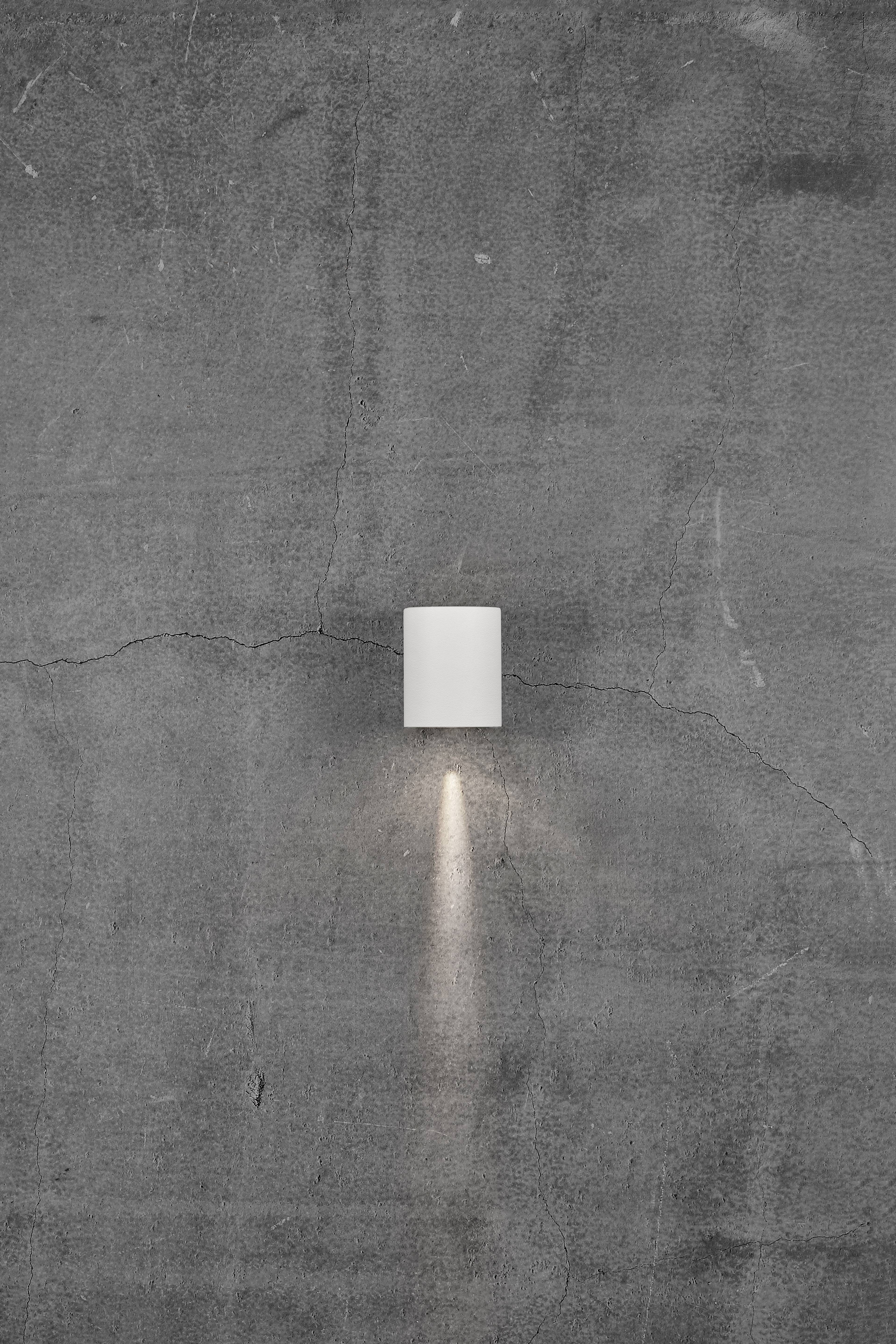 Wall light on a grey, textured wall, casting a narrow beam of light downwards. Minimalist design.
