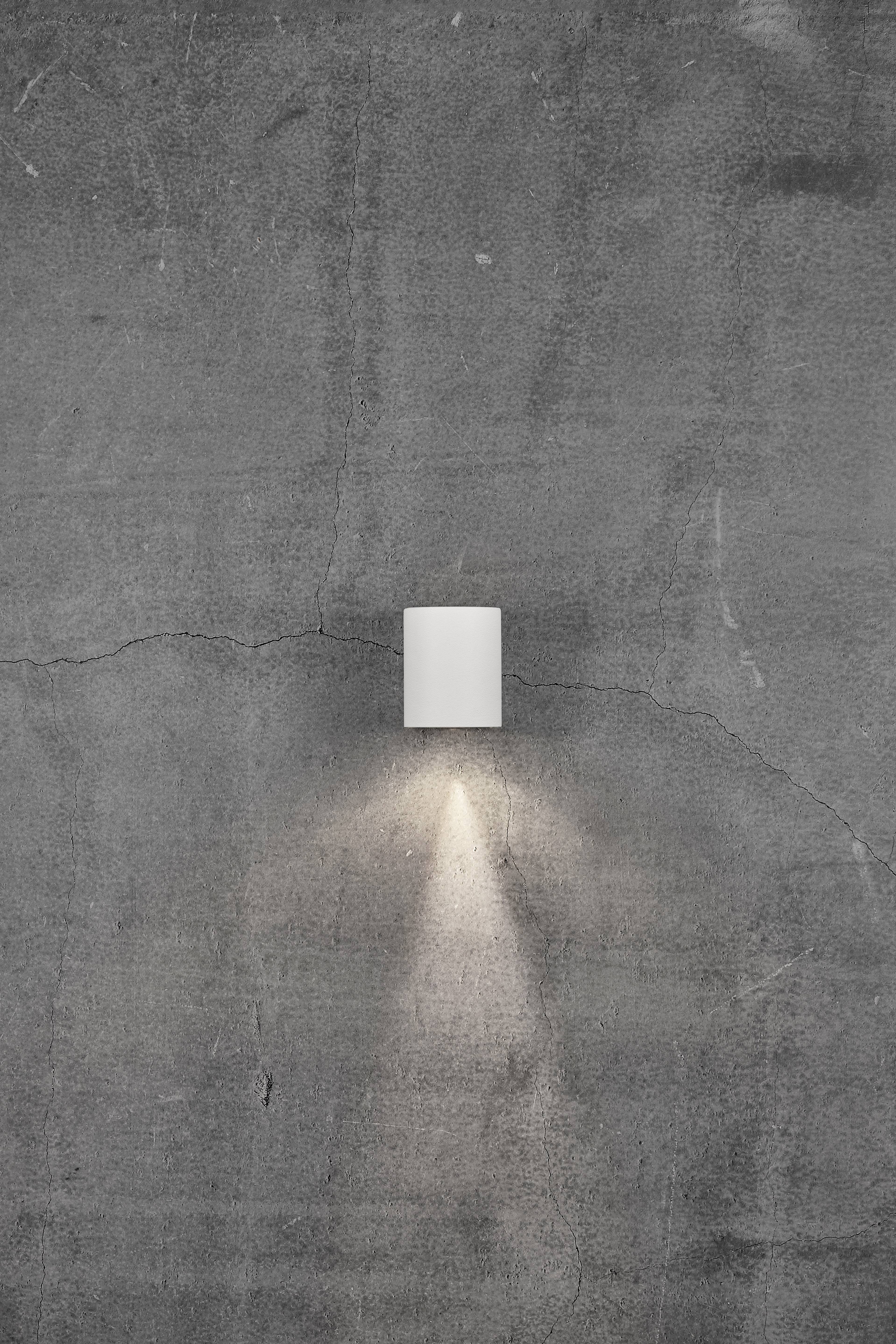 A minimalist wall light casts warm light onto a grey, cracked concrete wall. The light creates a soft, downward-directed glow.