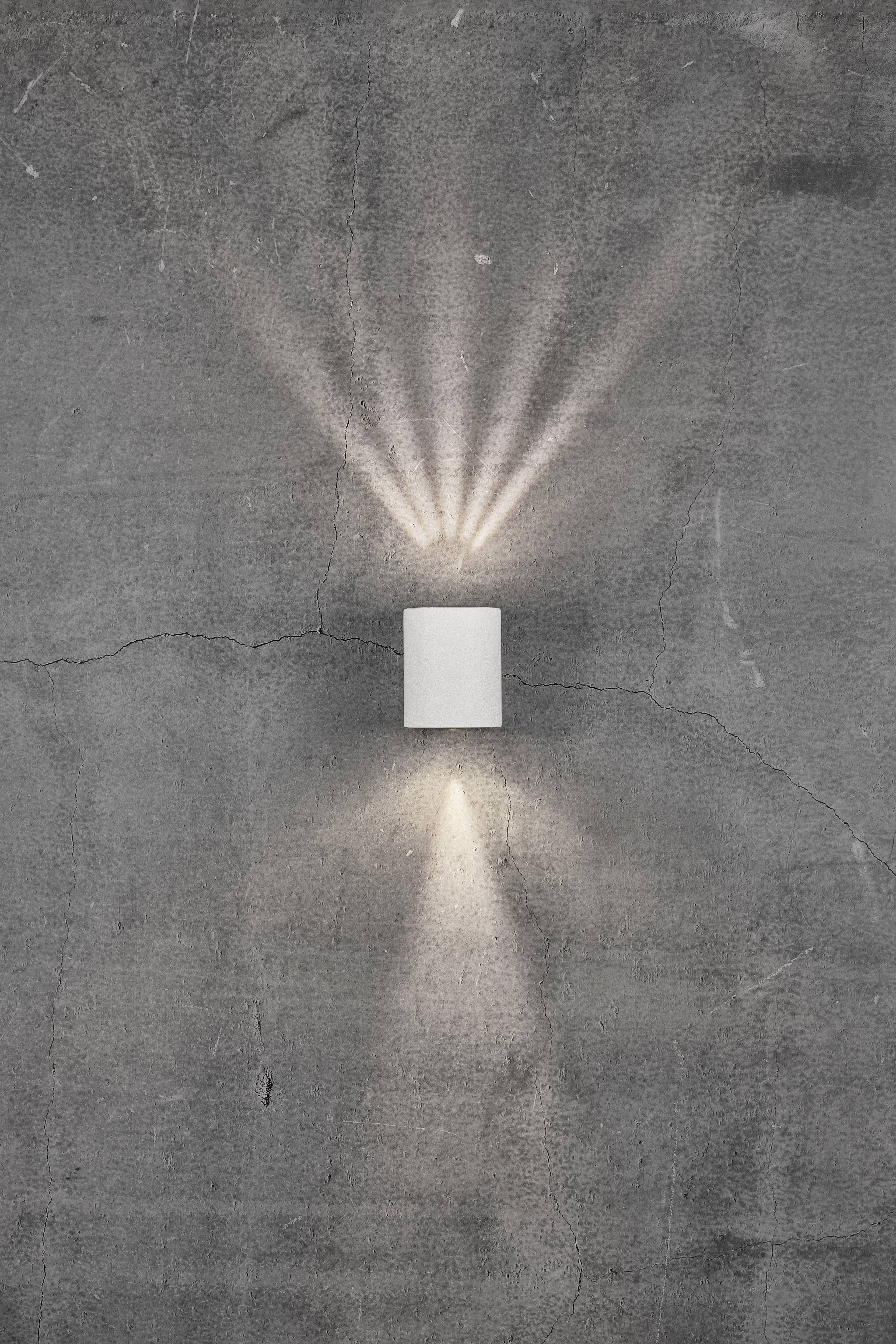 A cylindrical wall light on a grey concrete wall emits light rays upwards and downwards.
