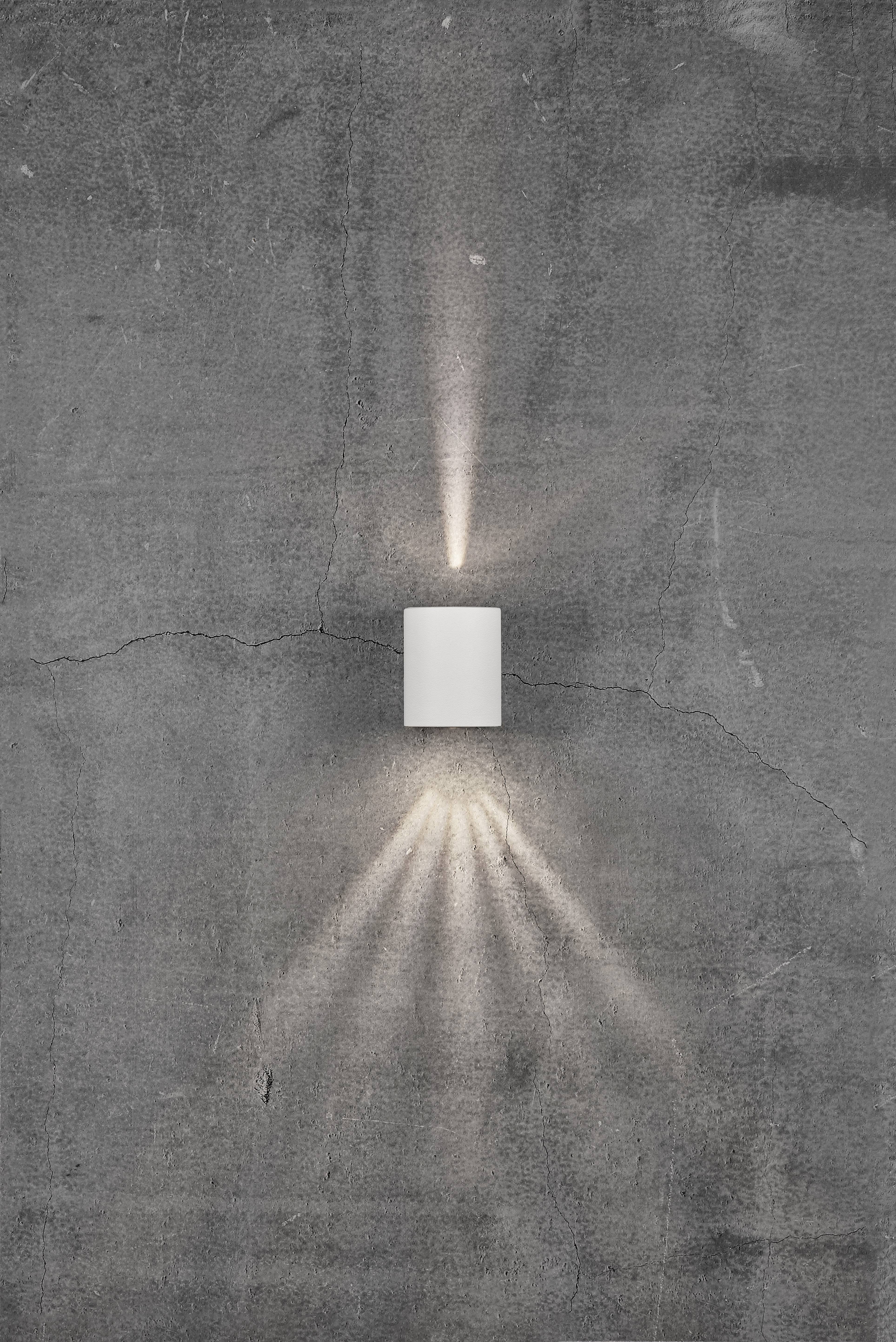 Wall light on a grey, textured concrete wall. The lamp illuminates upwards and downwards, creating ray-like light patterns.