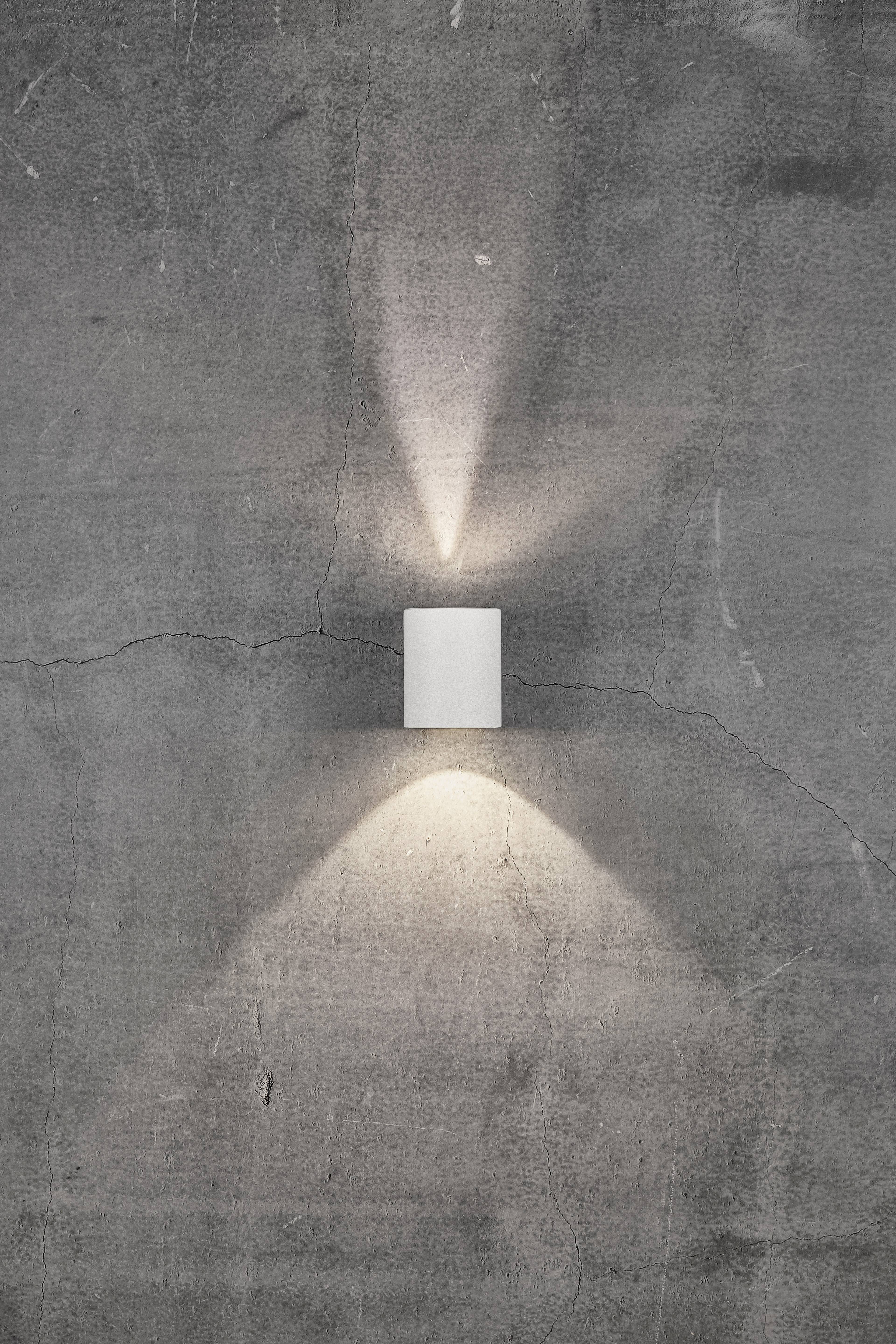 White wall lamp on a grey, cracked concrete wall, casting light upwards and downwards.