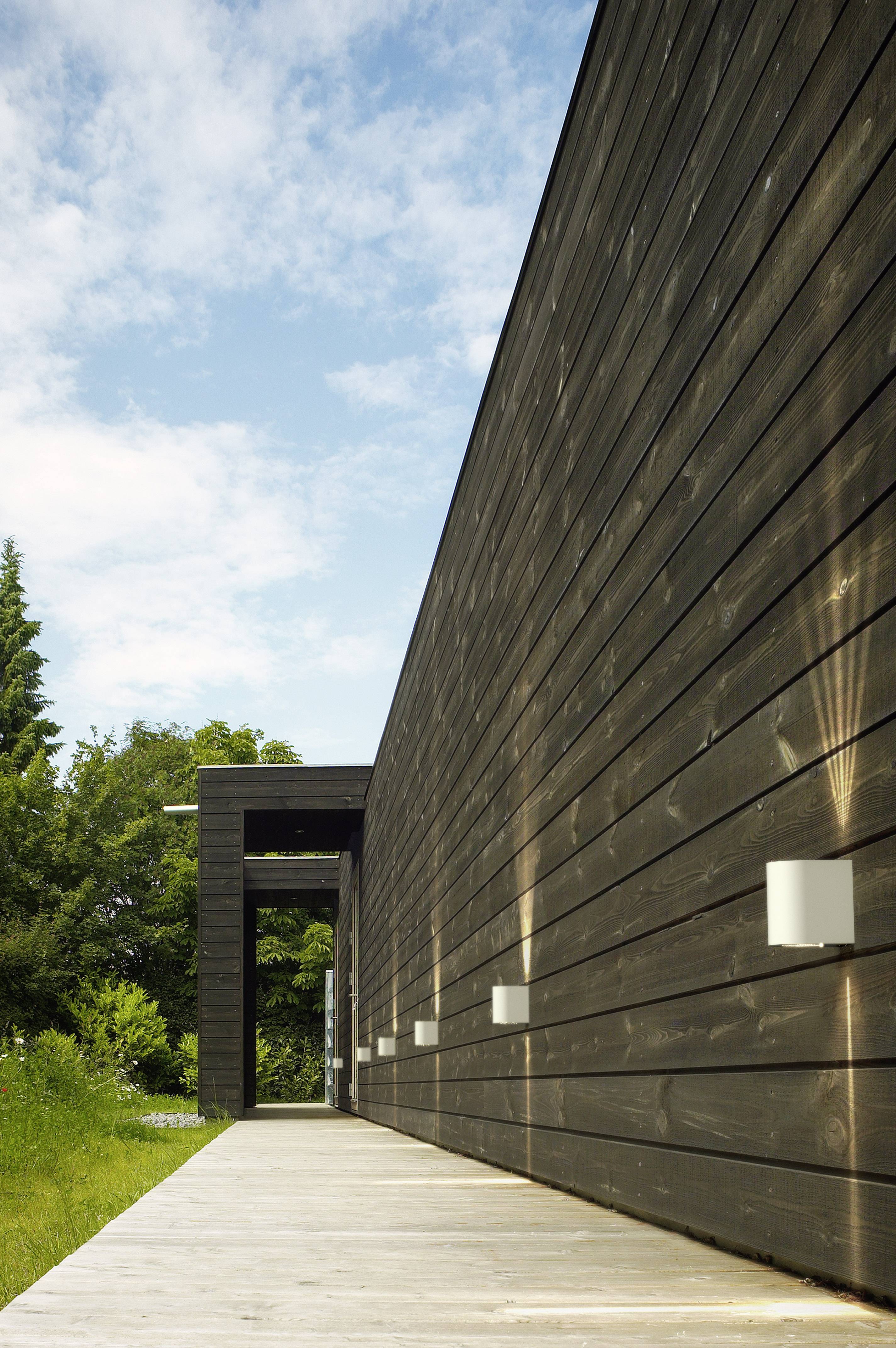 A modern wooden walkway runs alongside the dark wooden facade of a building. Sunlight illuminates the path.