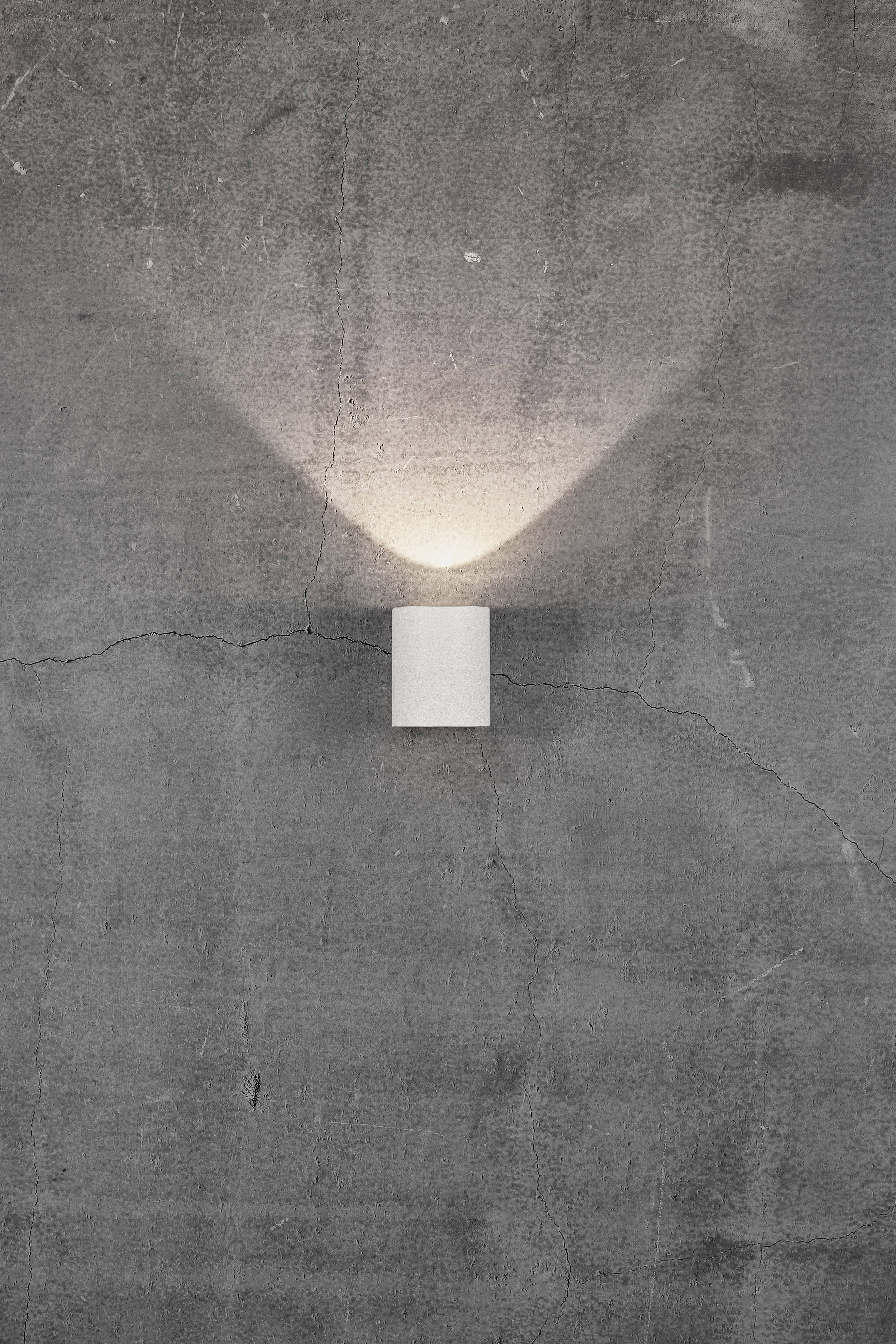 A simple white wall light gently illuminates a grey, cracked concrete wall, with the light radiating in a funnel-like shape upwards and downwards.